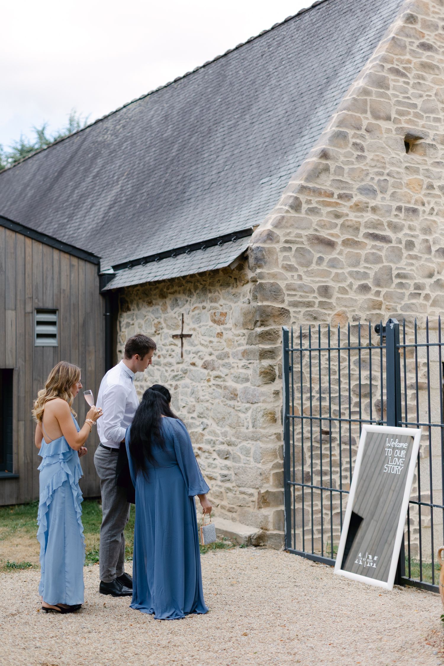 Trois invités au mariage examinant un panneau de bienvenue à l'extérieur d'un lieu de mariage en pierre rustique.