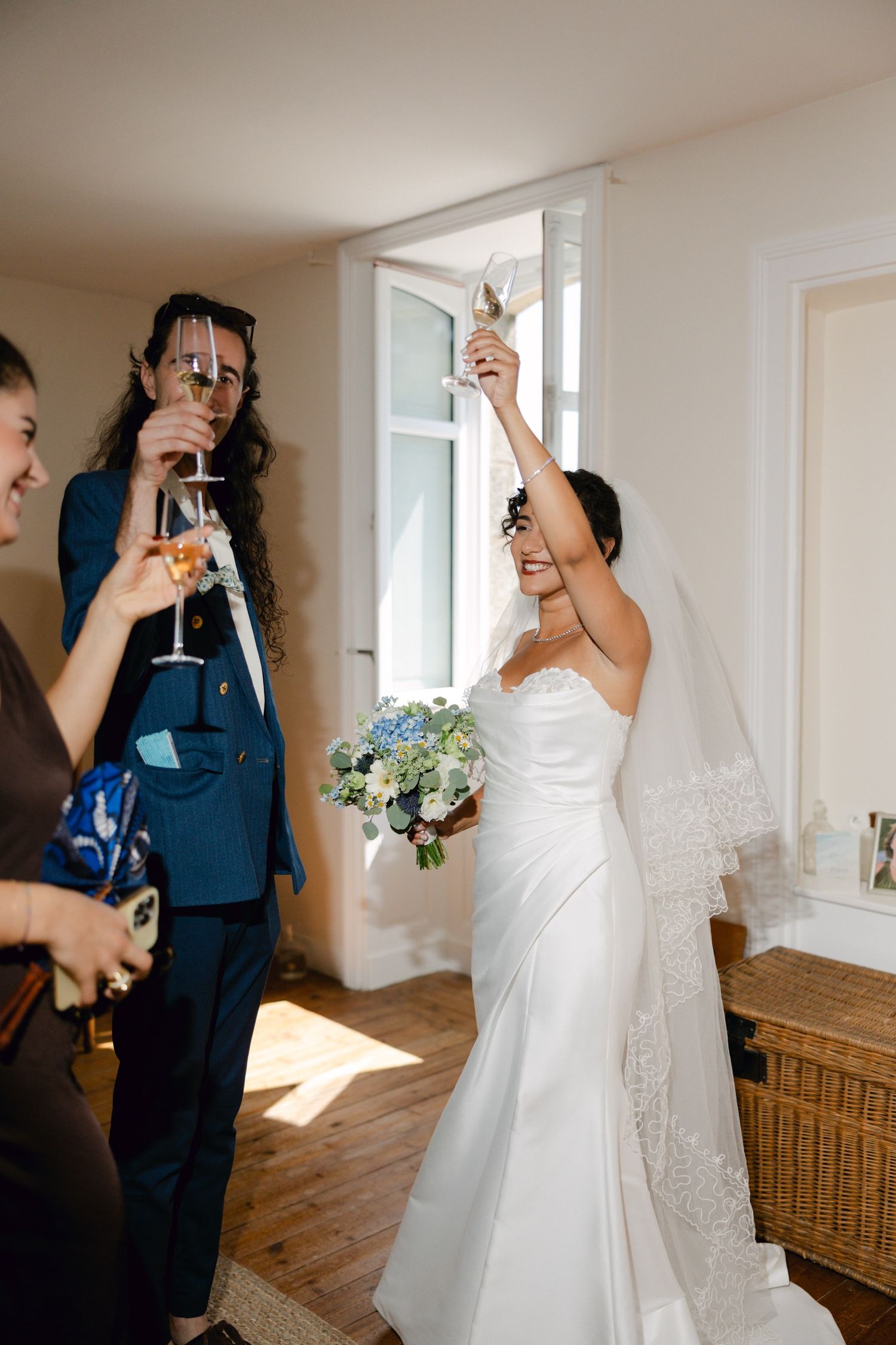 Bride in white gown raises champagne glass with guests during pre-wedding celebration indoors.