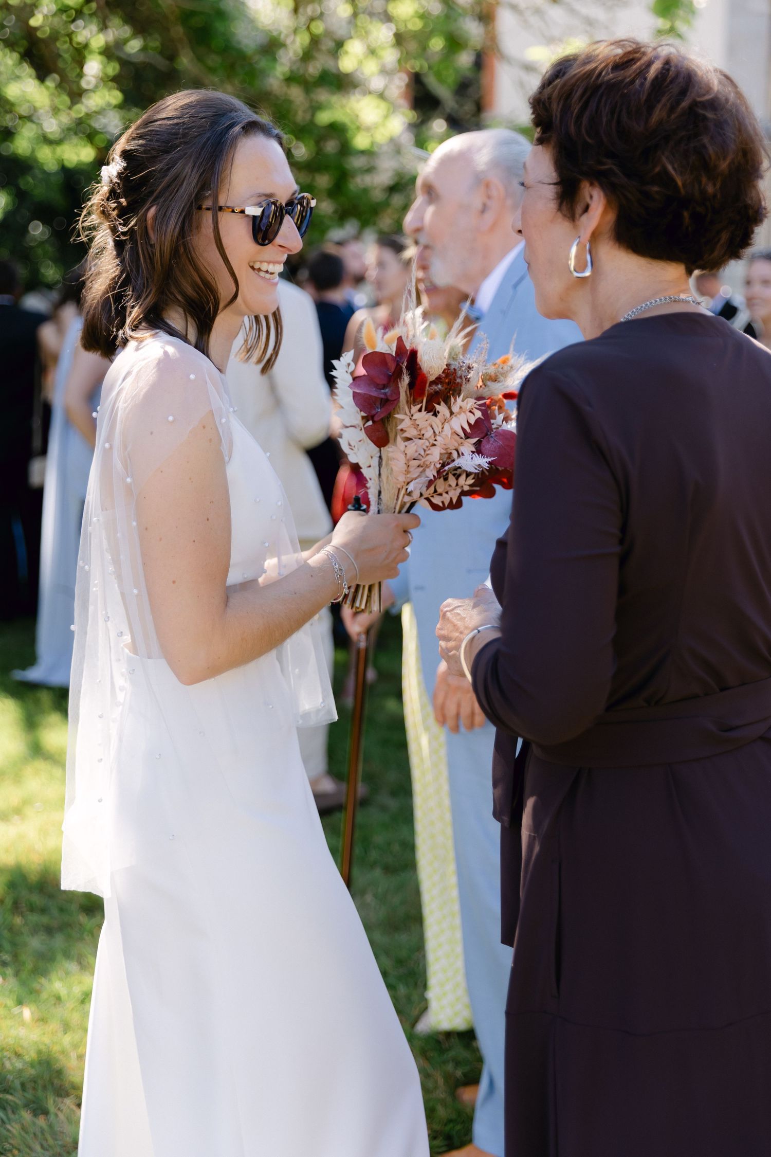 Mariée en robe blanche et lunettes de soleil tenant un bouquet, souriant à l'invité lors de la cérémonie de mariage en plein air.
