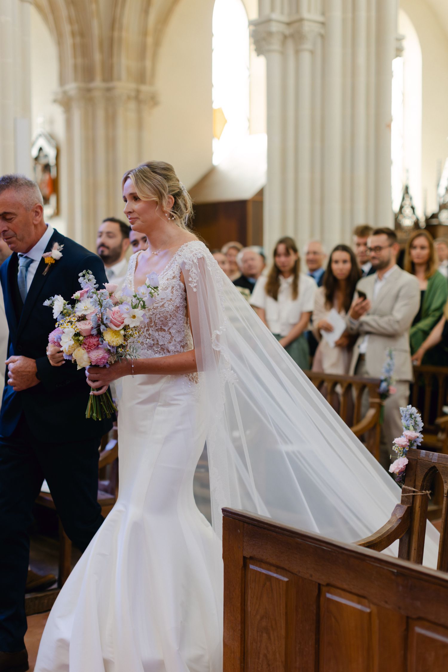 Mariée en robe de sirène en dentelle avec voile de cape fluide marche dans l'allée de l'église escortée par son père au mariage.