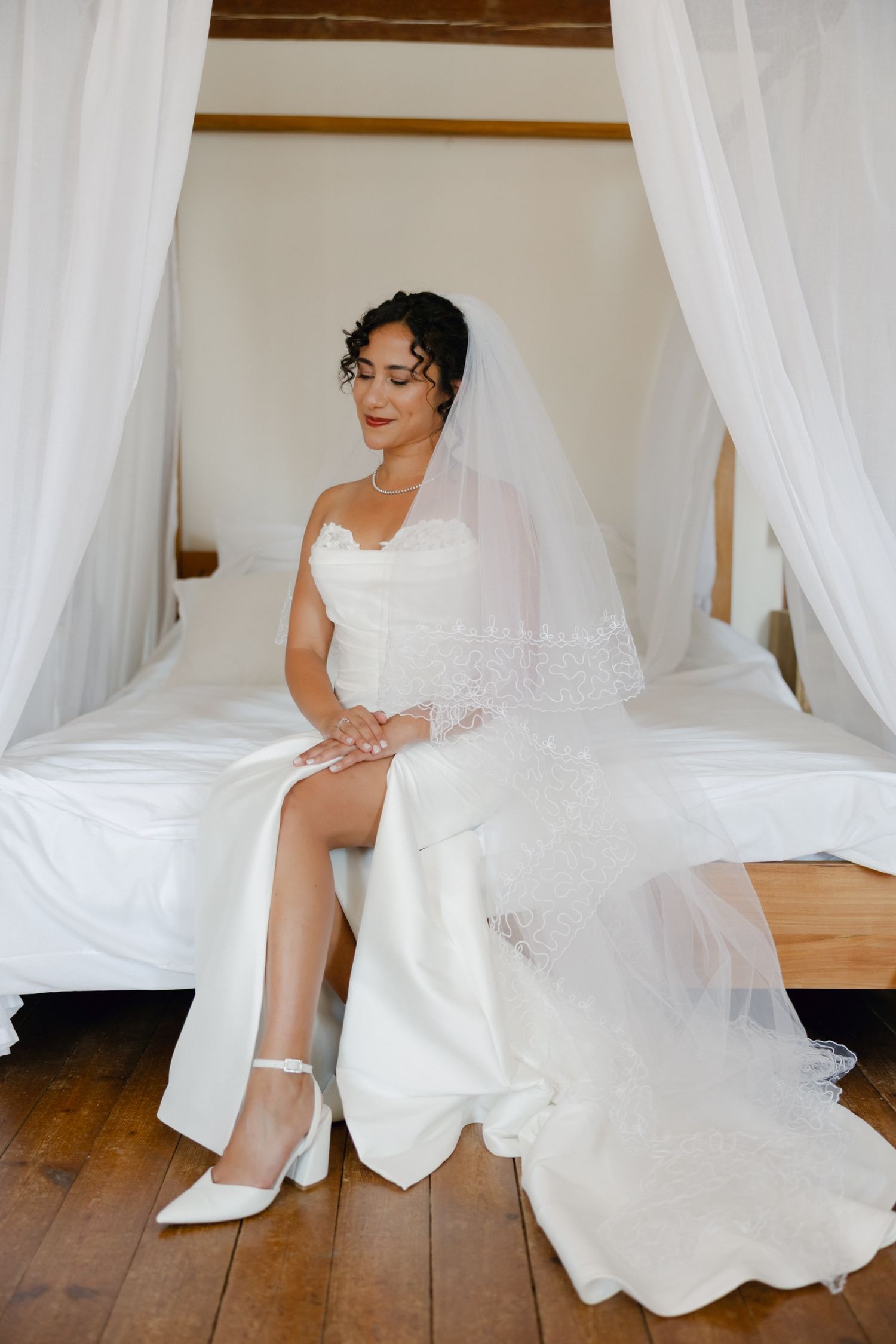 Bride in white strapless wedding gown and veil sits gracefully on a canopy bed with white curtains.