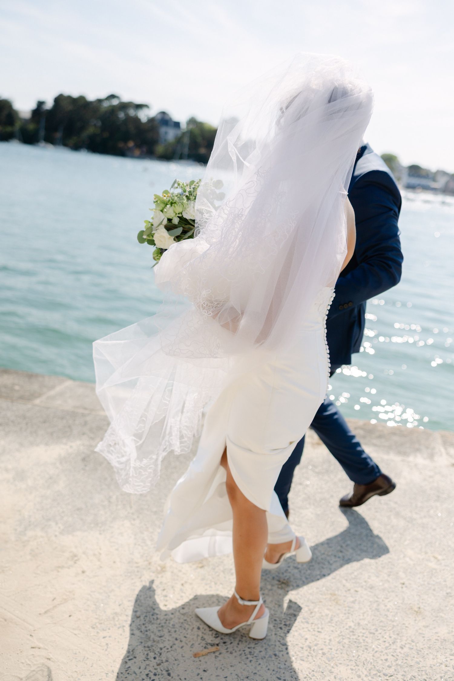 Bride in white dress and veil holding bouquet walks with groom in navy suit along waterfront on wedding day.