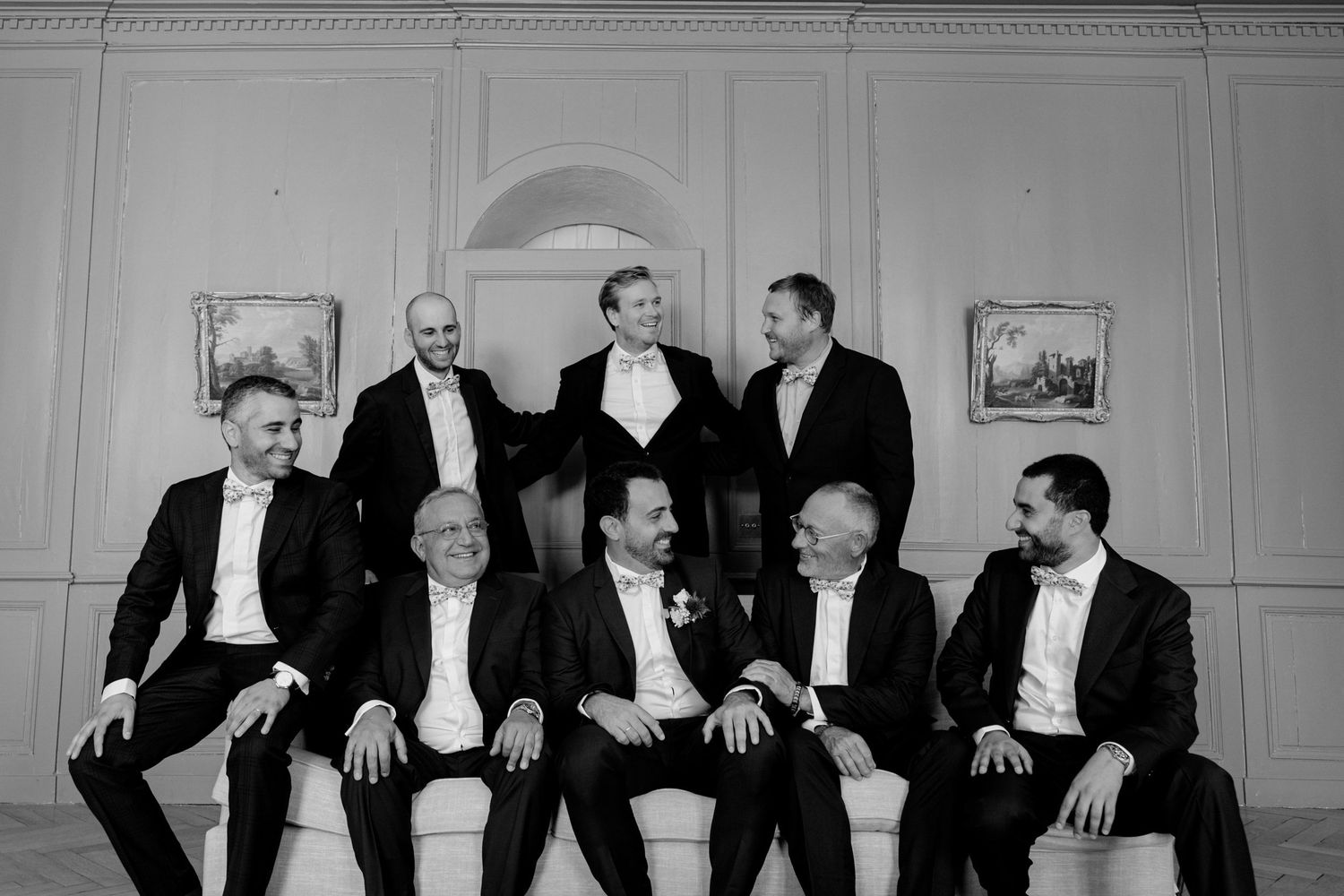 Black and white photo of nine groomsmen in suits posing together on a sofa in an elegant room.