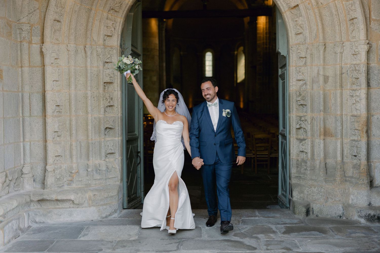 Joyful newlyweds exit stone church, bride in white gown raising bouquet, groom in navy suit, holding hands.