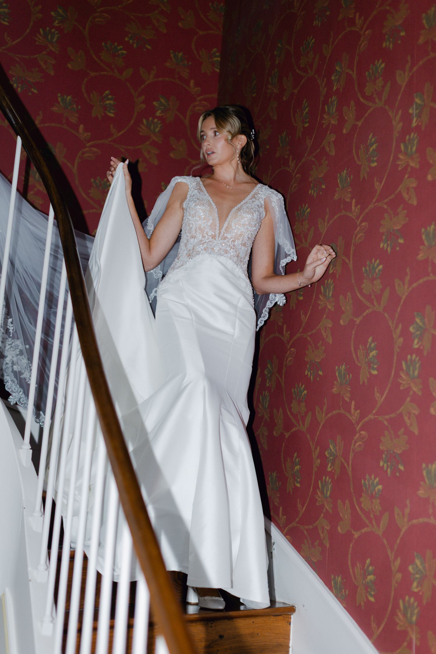 La mariée en robe de mariée en dentelle blanche ajustée pose dans l'escalier contre du papier peint floral rouge tenant sa cape.