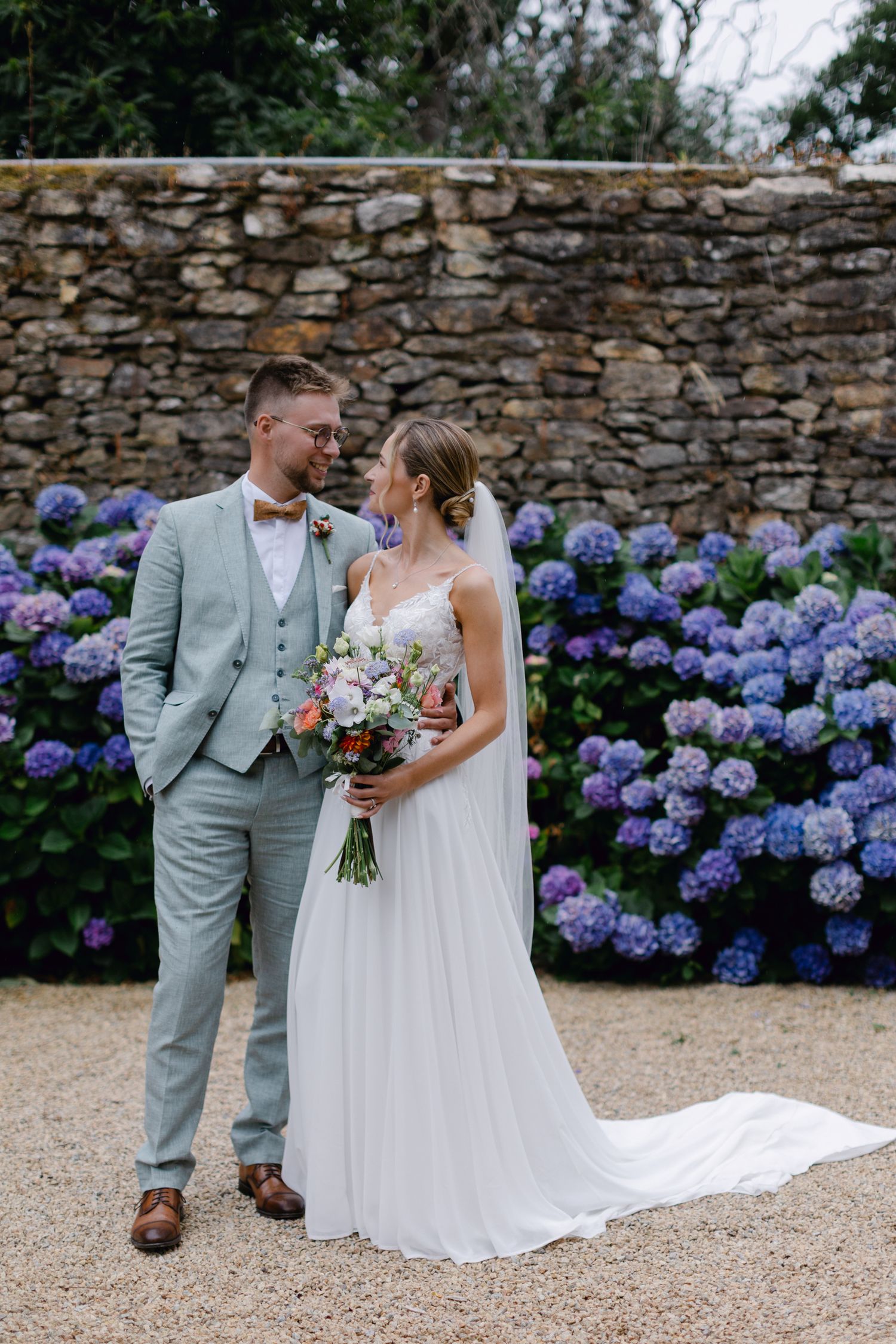 La mariée et le marié se regardant devant un mur de pierre avec des hortensias violets en fleurs.
