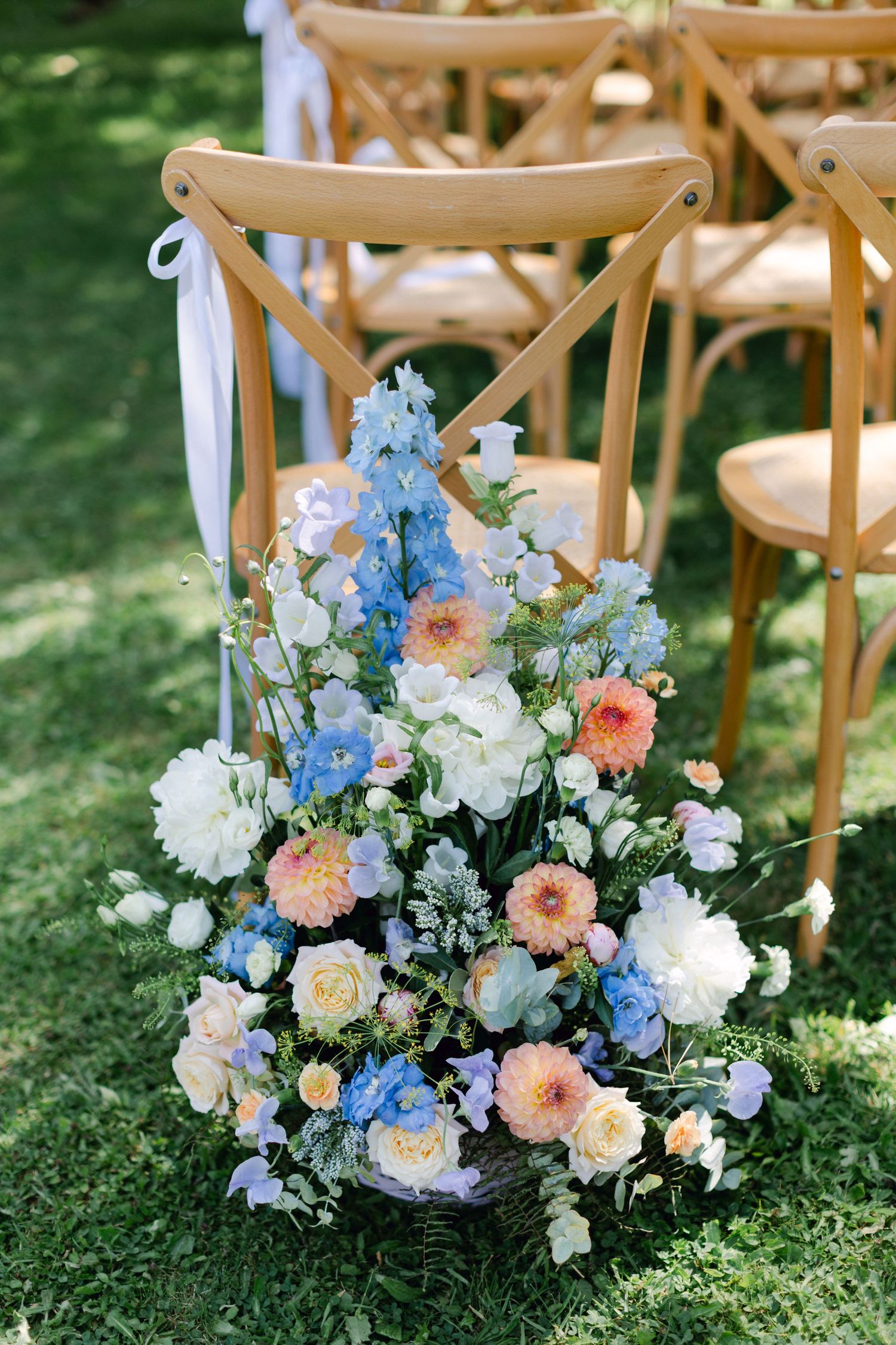Arrangement floral luxuriant de fleurs bleues, pêches et blanches dans un panier à côté de chaises en bois lors d'une cérémonie en plein air