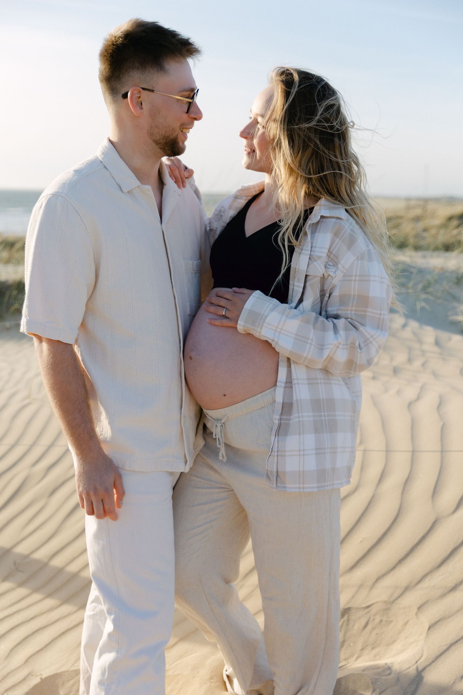 Couple enceinte en tenue décontractée souriant l'un à l'autre sur les dunes de la plage de sable sous la chaude lumière du soleil.