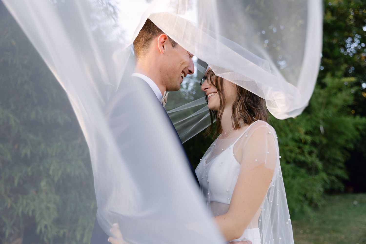 La mariée et le marié souriant sous un voile blanc fluide à l'extérieur le jour de leur mariage.