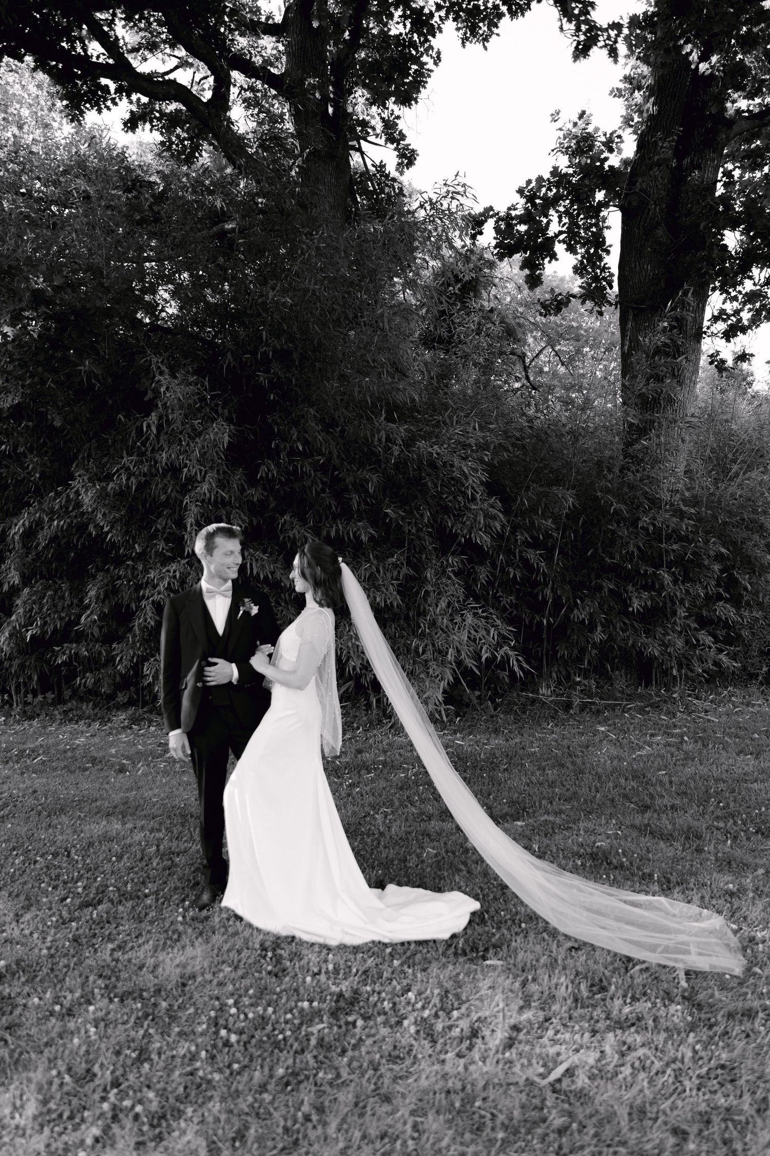Photo en noir et blanc de la mariée et du marié debout sur l'herbe avec un voile de cathédrale fluide et de grands arbres en arrière-plan.