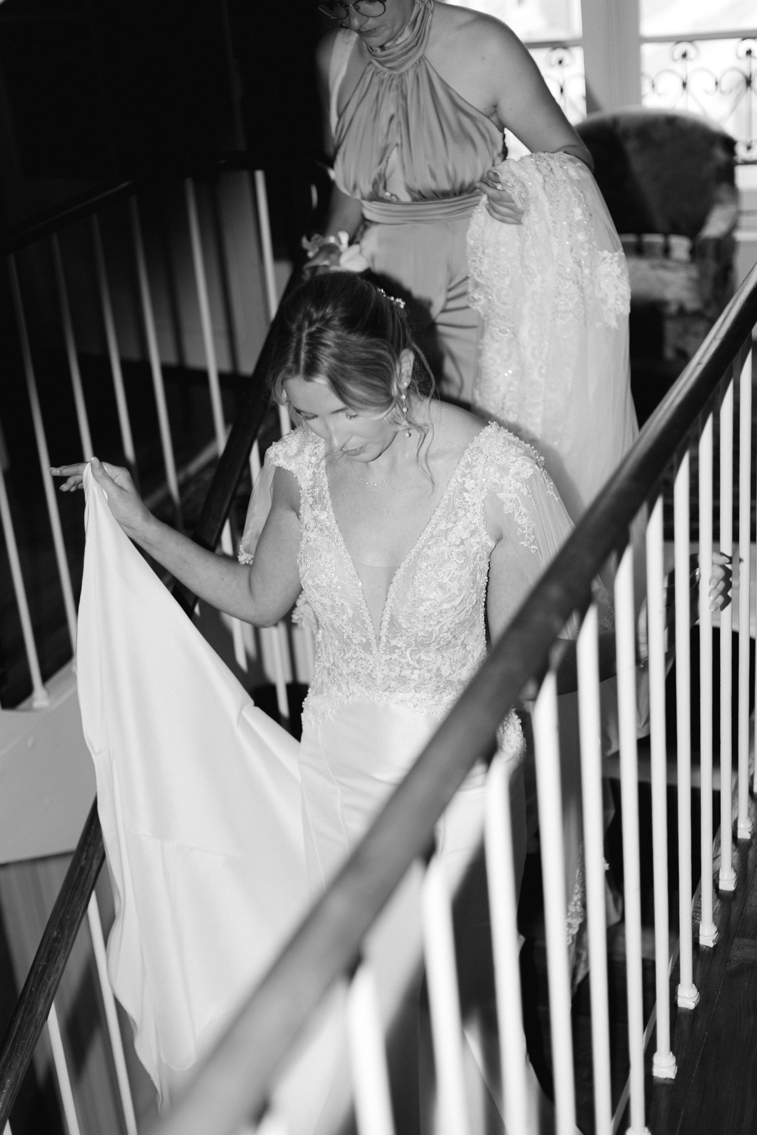 Photo en noir et blanc d'une mariée émotive en robe de mariée en dentelle sur l'escalier avec demoiselle d'honneur derrière elle.