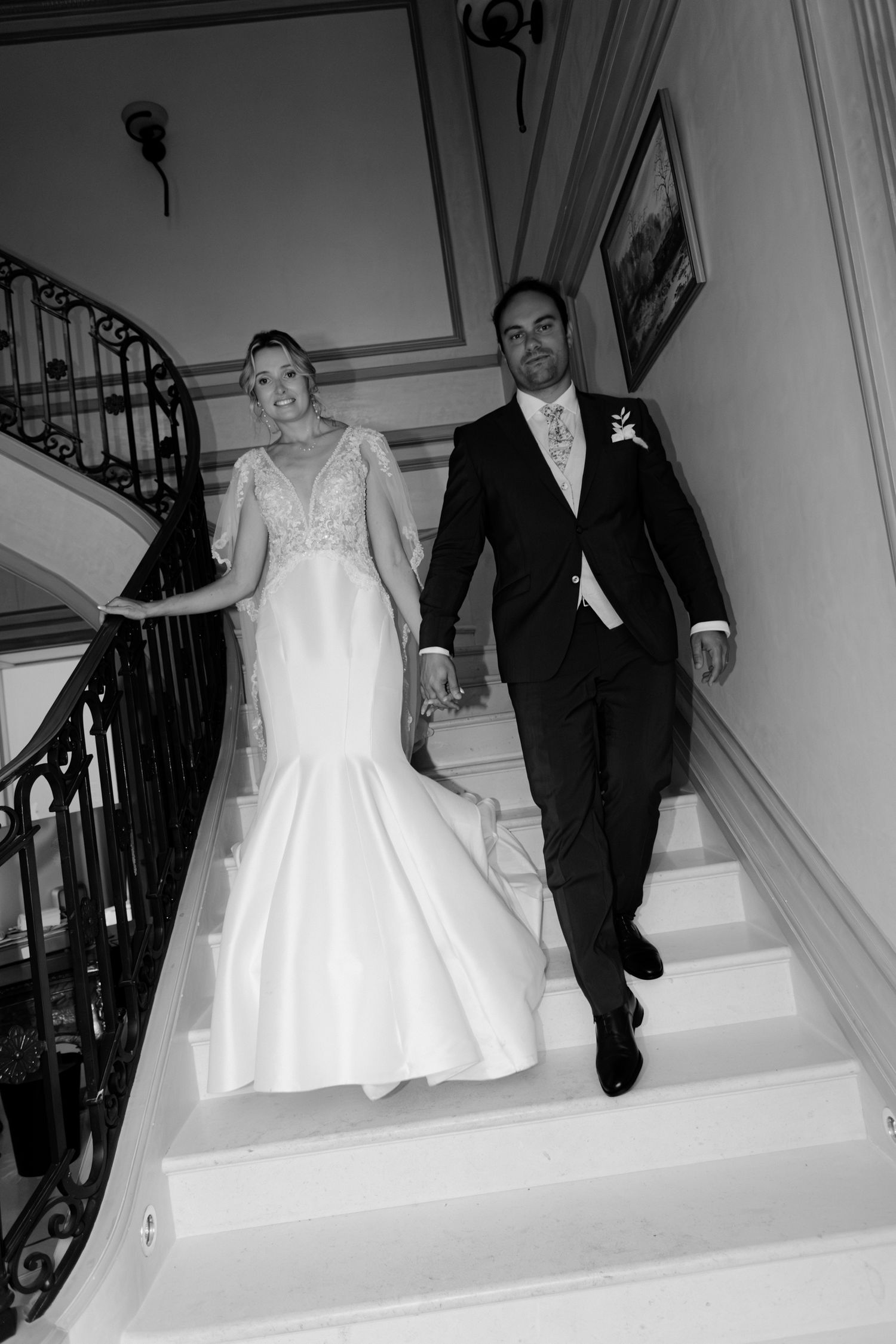 Photo en noir et blanc de la mariée et du marié marchant main dans la main dans l'escalier orné le jour du mariage.