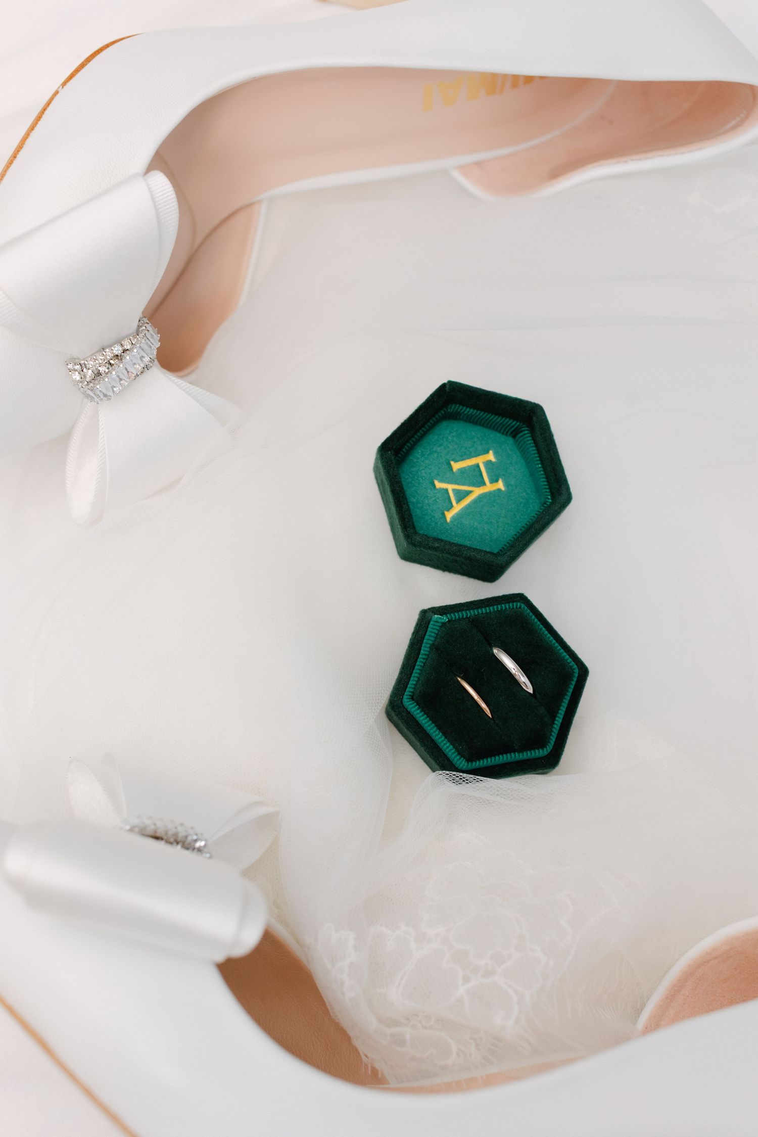 Alliances en or dans une boîte à bague hexagonale en velours vert à côté de talons de mariée en satin blanc avec détail d'arc en cristal.
