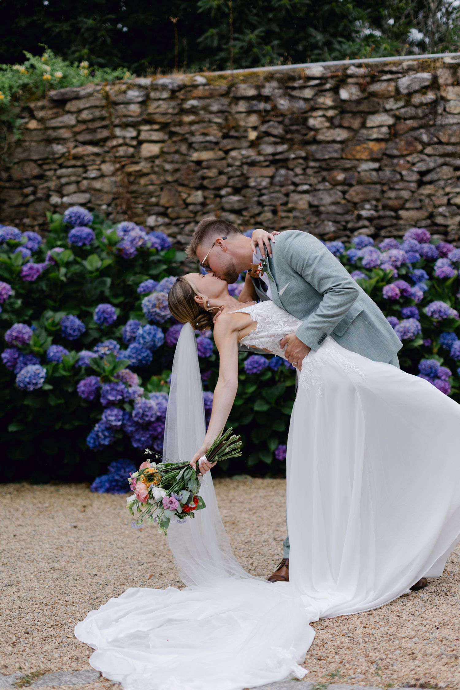 Le marié trempant la mariée pour un baiser devant un mur d'hortensias violets vibrants dans un lieu extérieur.