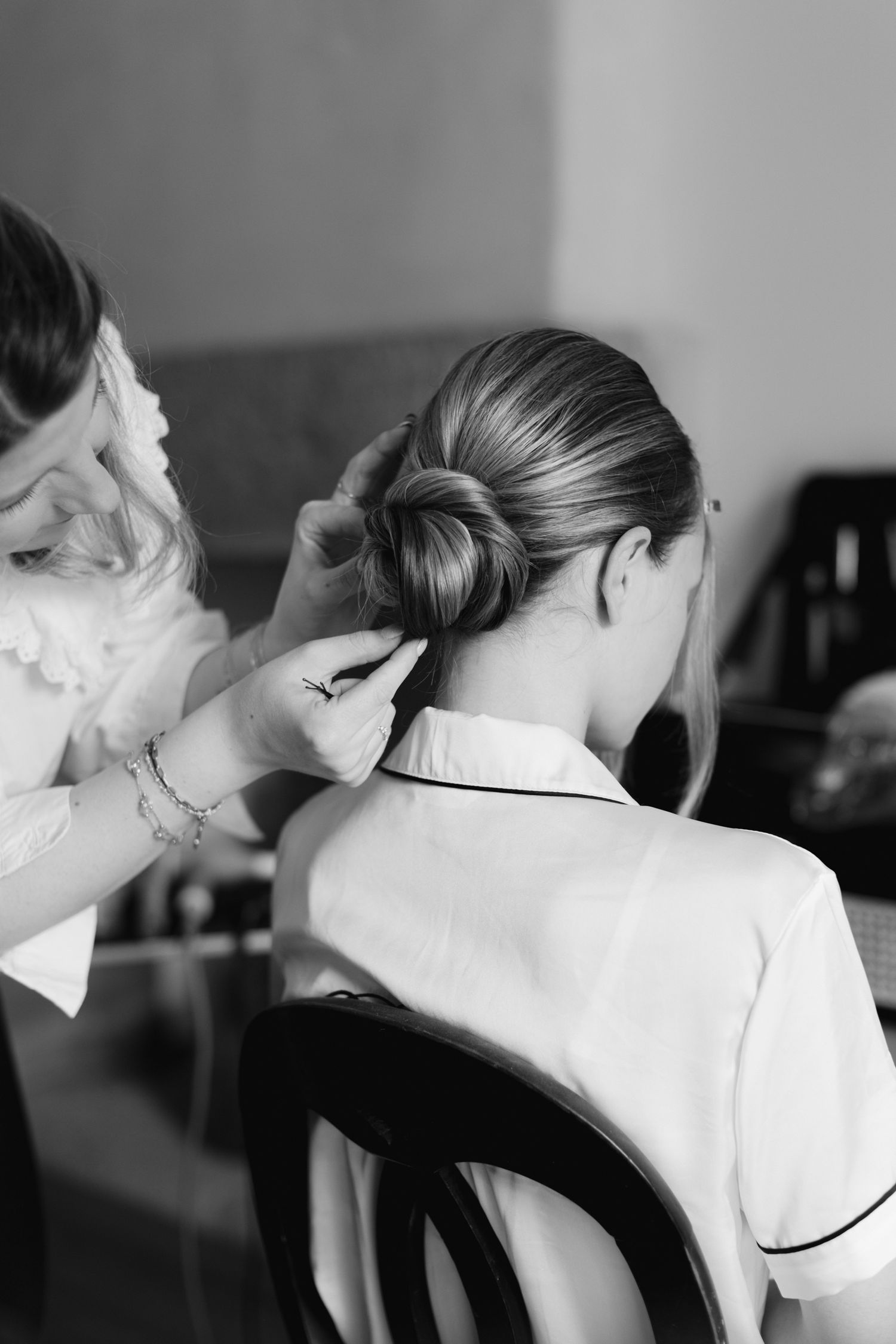 Photo en noir et blanc d'un coiffeur épinglant un chignon bas élégant sur une mariée assise.