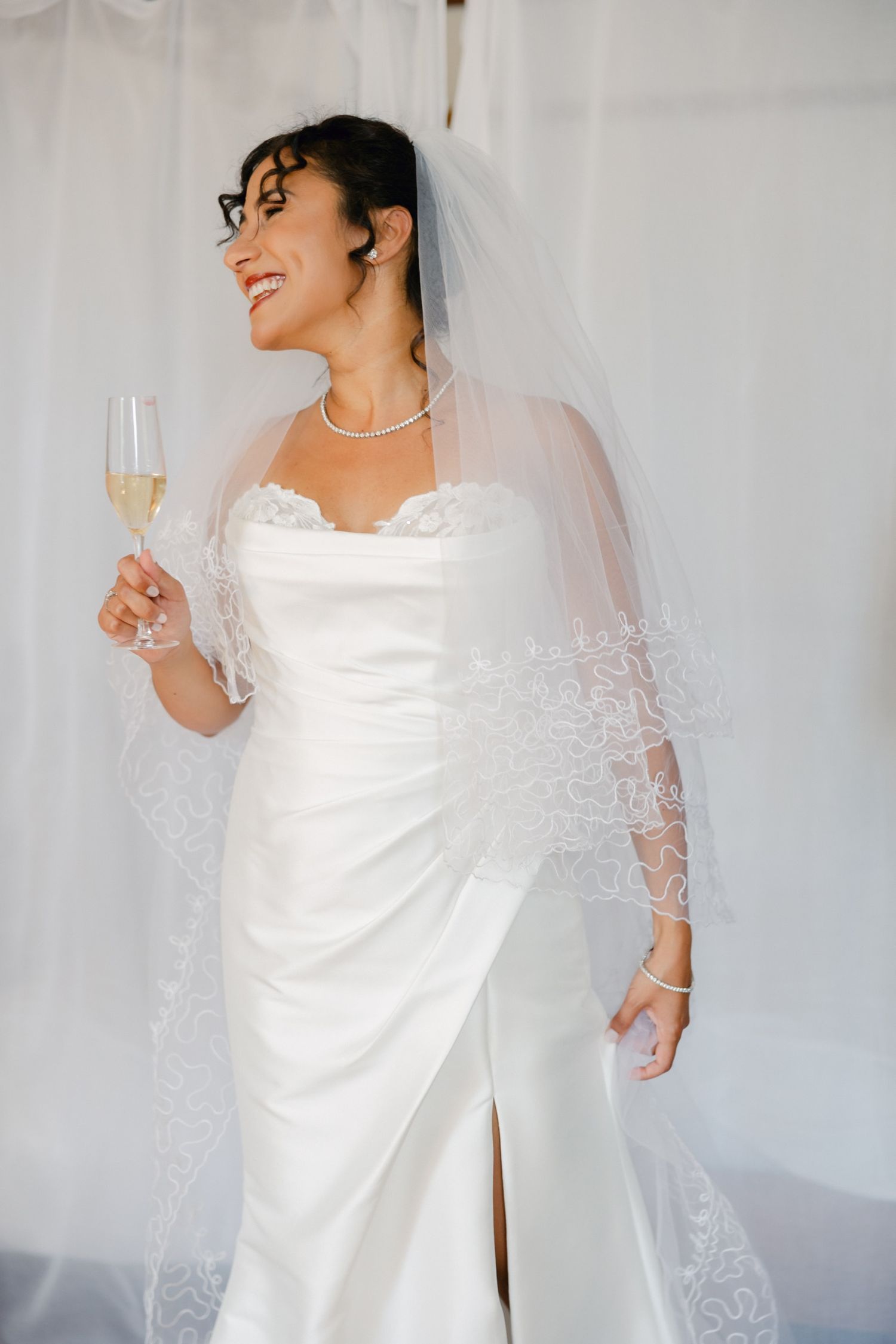 Smiling bride in white strapless gown and lace veil holds champagne glass, wearing pearl necklace and bracelet.