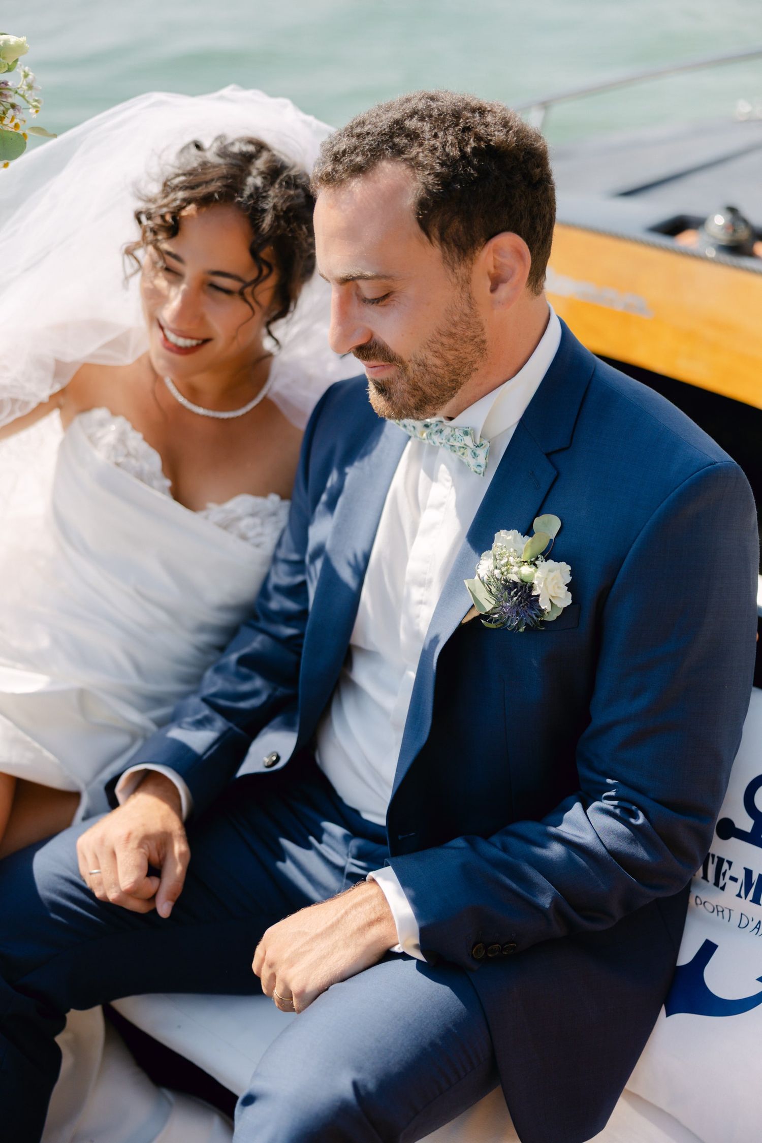 Bride and groom sitting on a boat, groom in navy suit with floral boutonniere, bride in white gown smiling warmly.