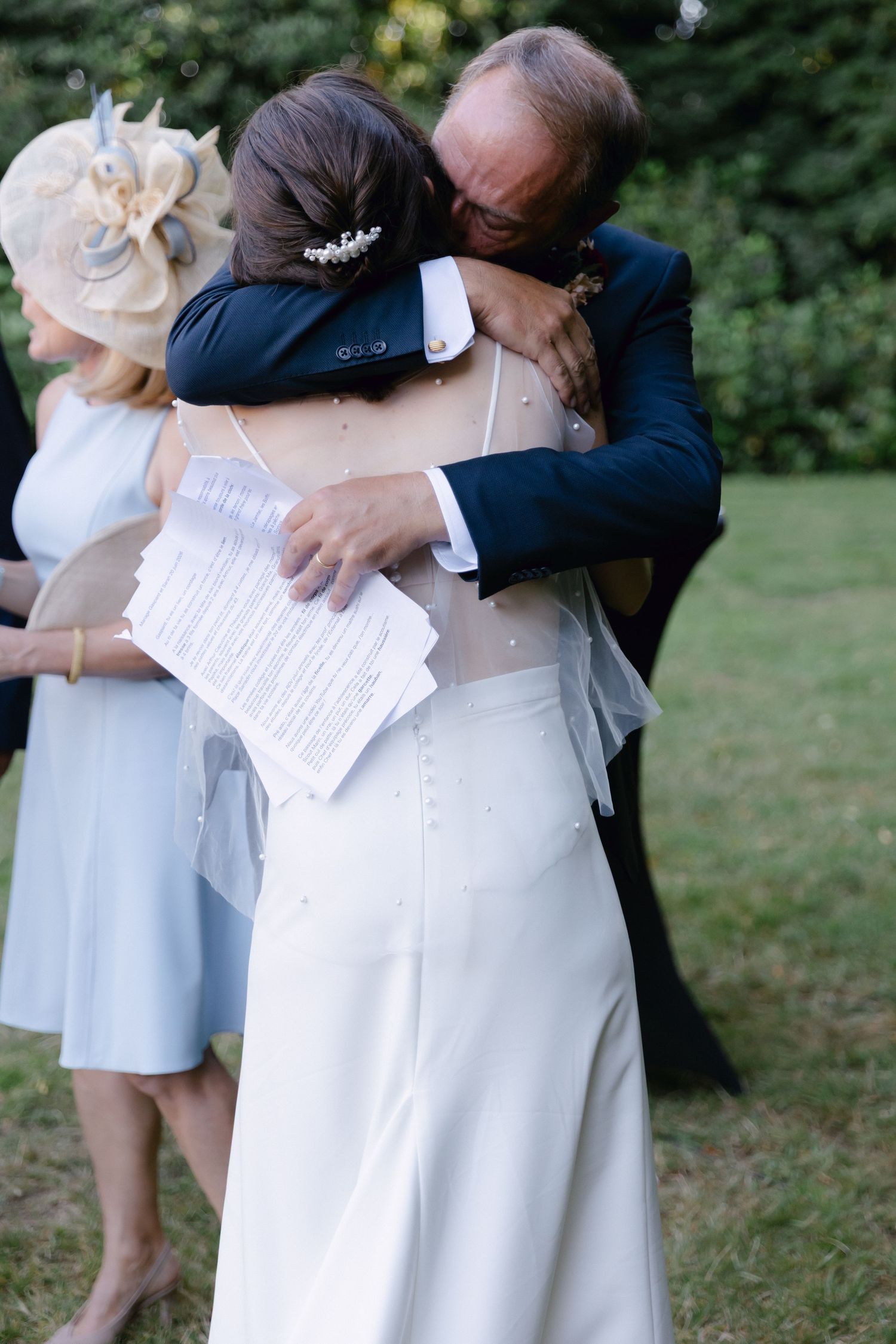 Mariée en robe blanche embrassant un homme plus âgé en costume bleu marine lors d'une cérémonie de mariage en plein air, avec une femme en r