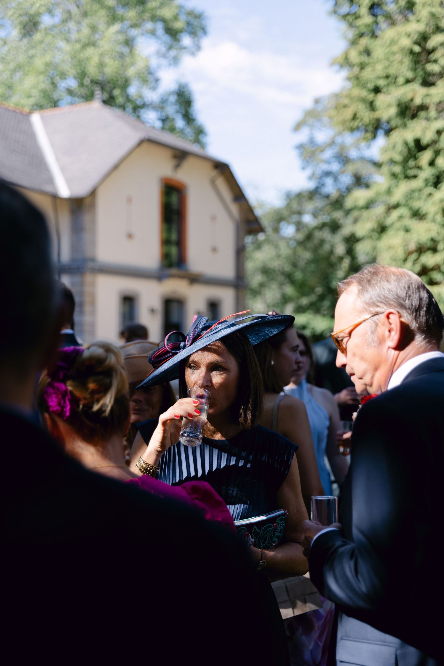 Femme élégamment habillée avec un chapeau de fascinateur bleu marine sirotant un verre lors d'une fête de jardin en plein air.