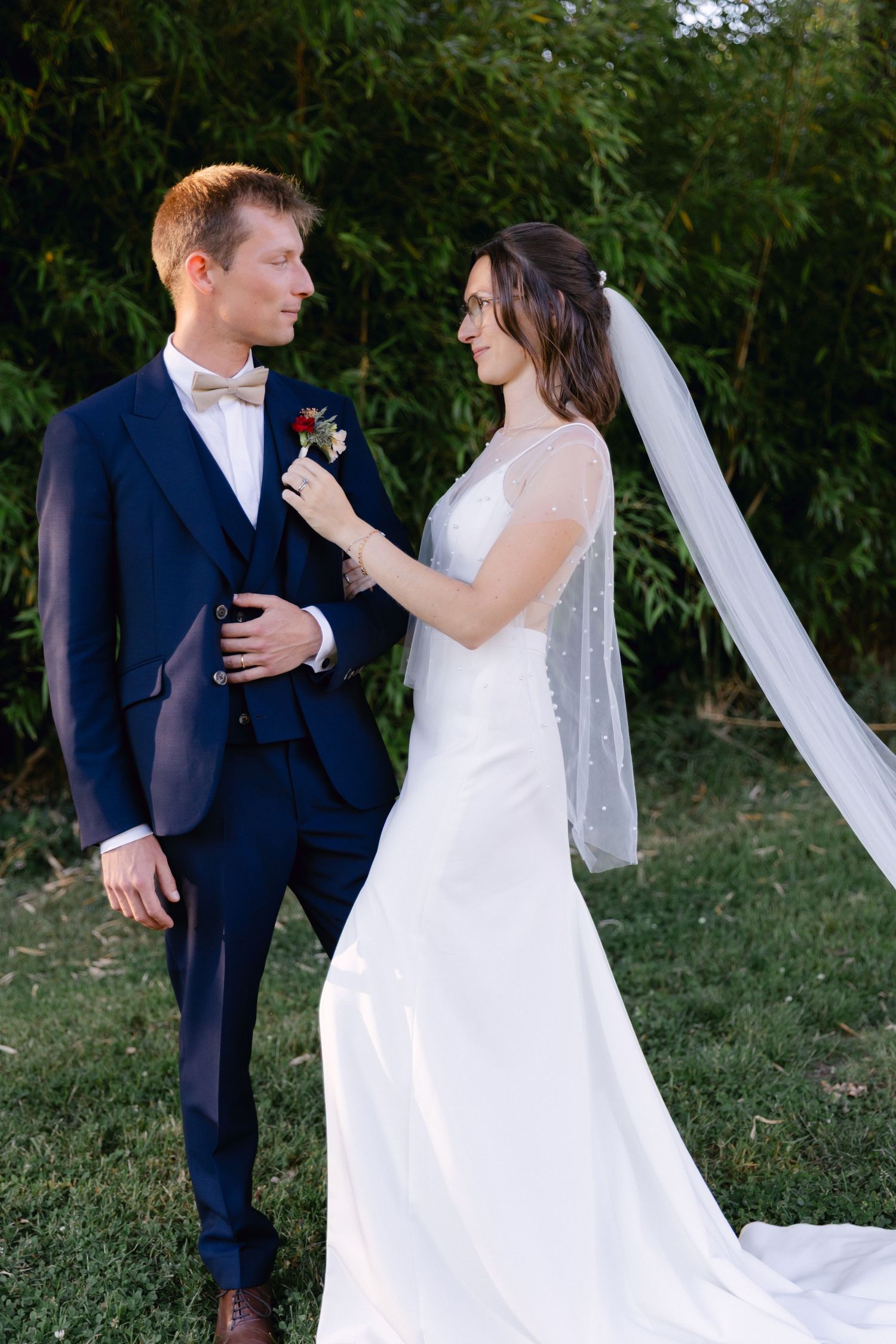 La mariée en robe blanche ajuste la boutonnière du marié alors qu'ils partagent un moment tendre à l'extérieur le jour de leur mariage.