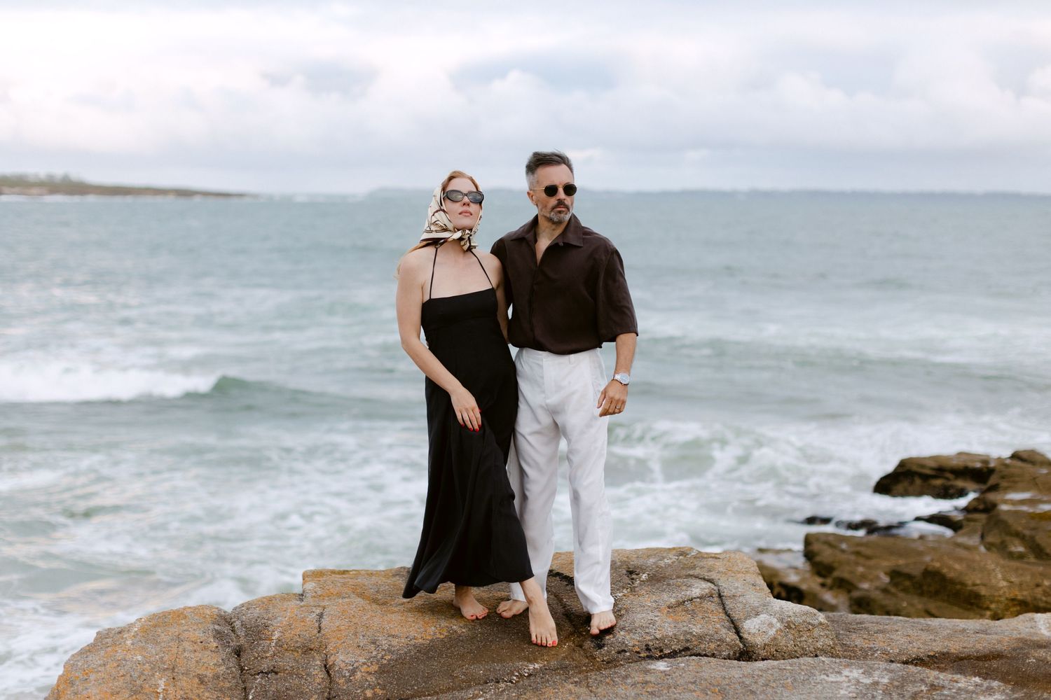 Couple posant sur des rochers côtiers avec les vagues de l'océan derrière eux, femme en robe noire et homme en pantalon blanc.