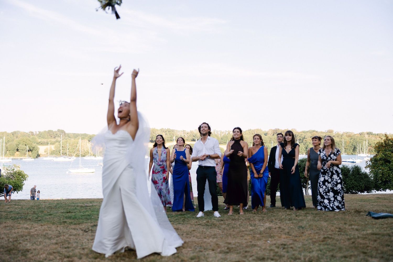Bride tossing bouquet to guests outdoors at waterfront wedding reception on sunny day.