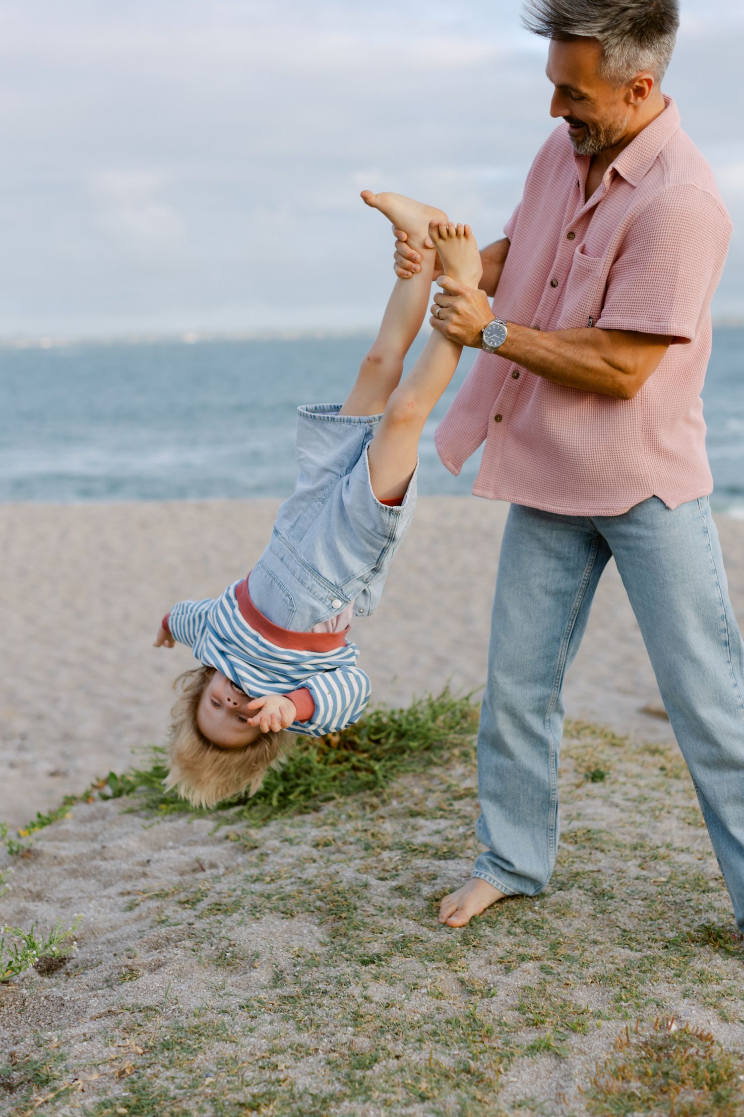 Homme tenant un enfant à l'envers sur la plage de sable, jouant et riant ensemble près du rivage de l'océan.