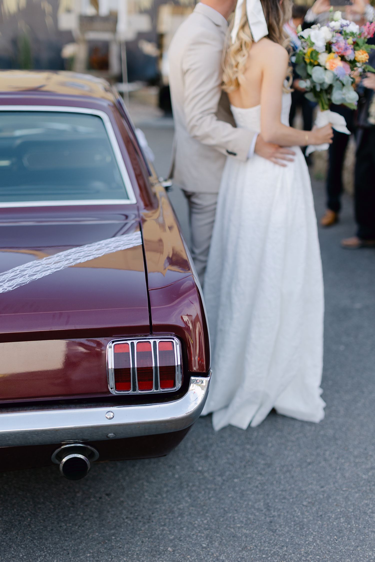 La mariée en robe blanche et le marié en costume beige se tiennent à côté d'une voiture de mariage Ford Mustang bordeaux classique.