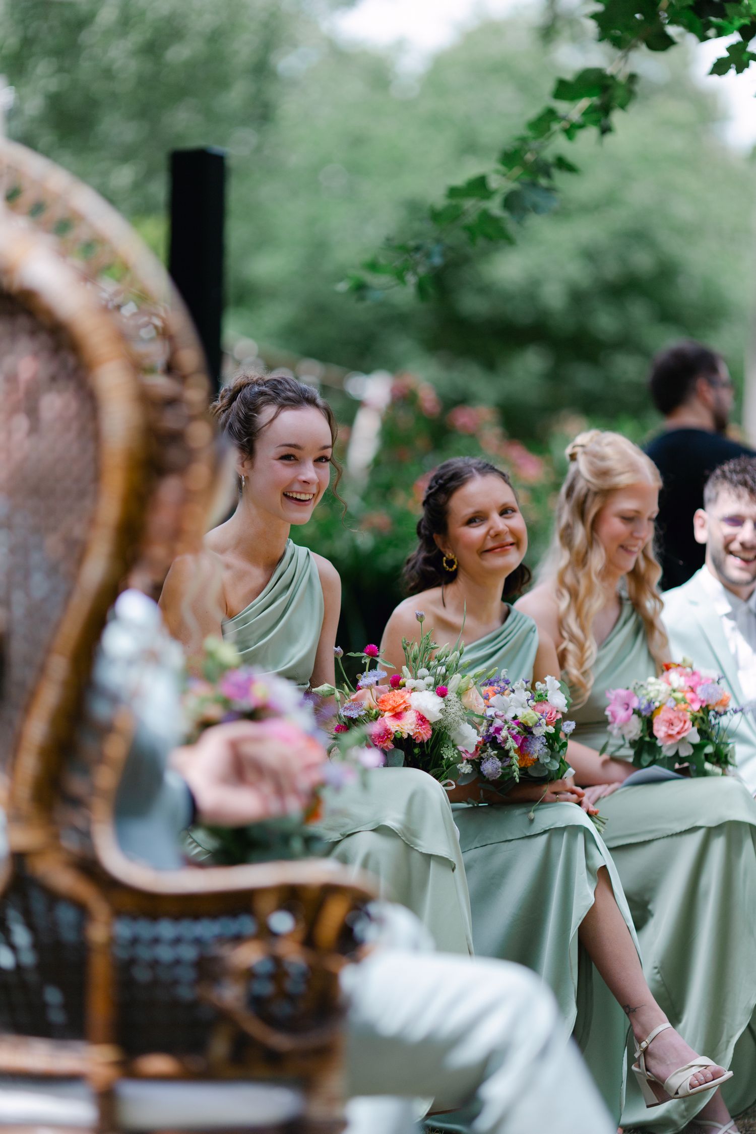 Les demoiselles d'honneur en robes vert sauge sourient tout en tenant des bouquets colorés lors d'un mariage en plein air.