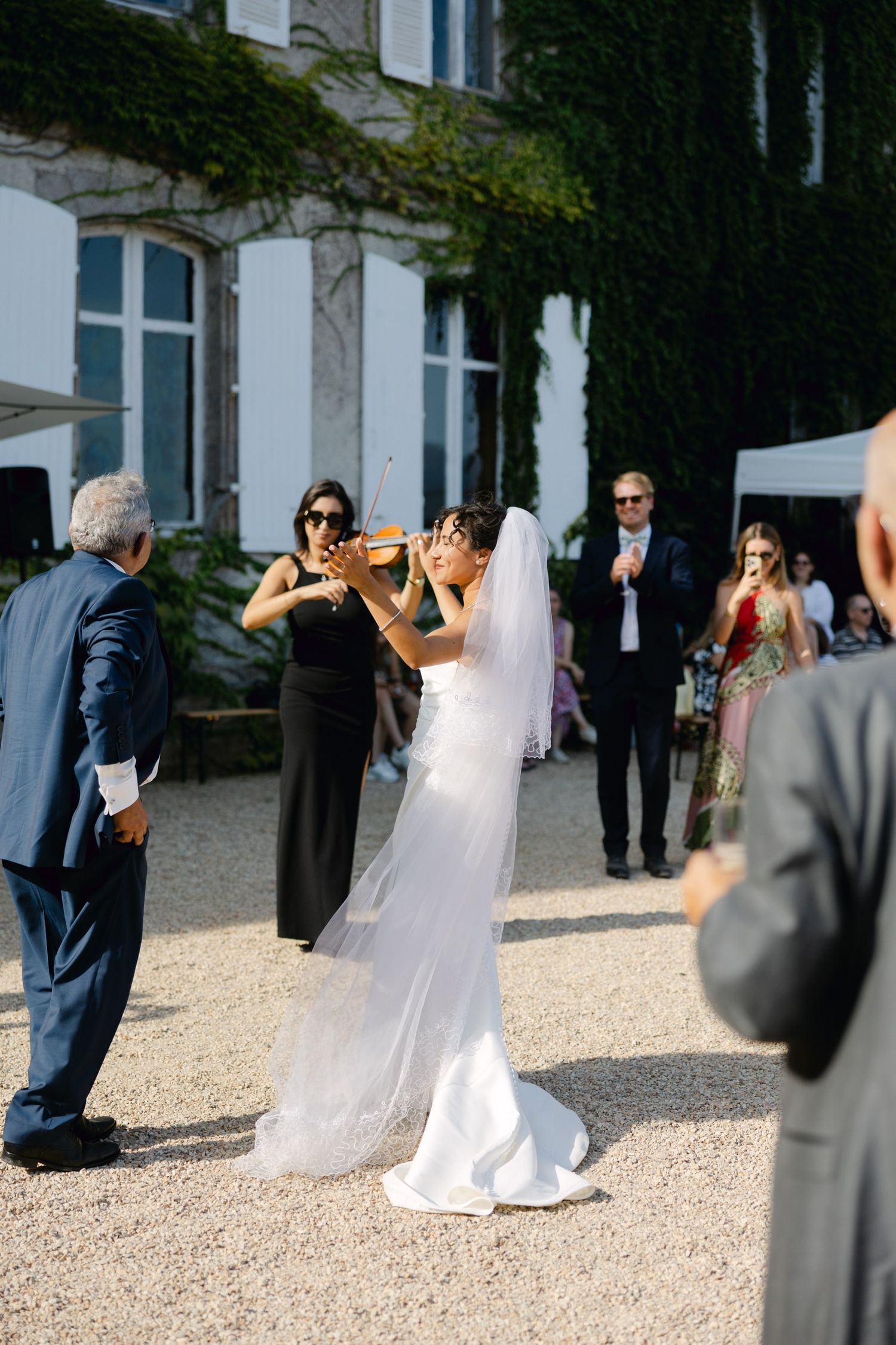 Bride in white gown plays violin outdoors at elegant chateau wedding reception with guests watching.