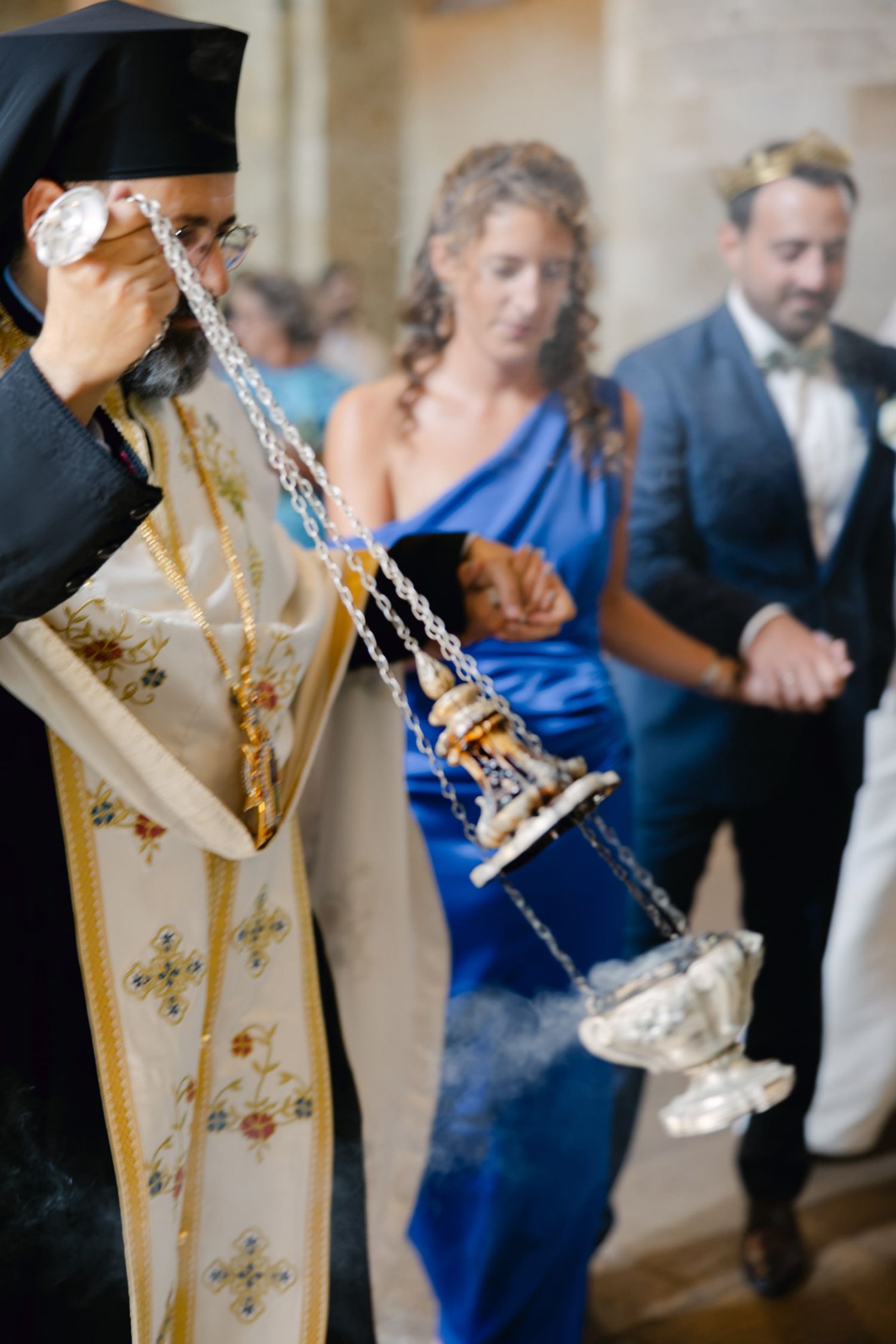 Orthodox priest swinging a censer with incense during a religious ceremony, with guests in formal attire behind him.