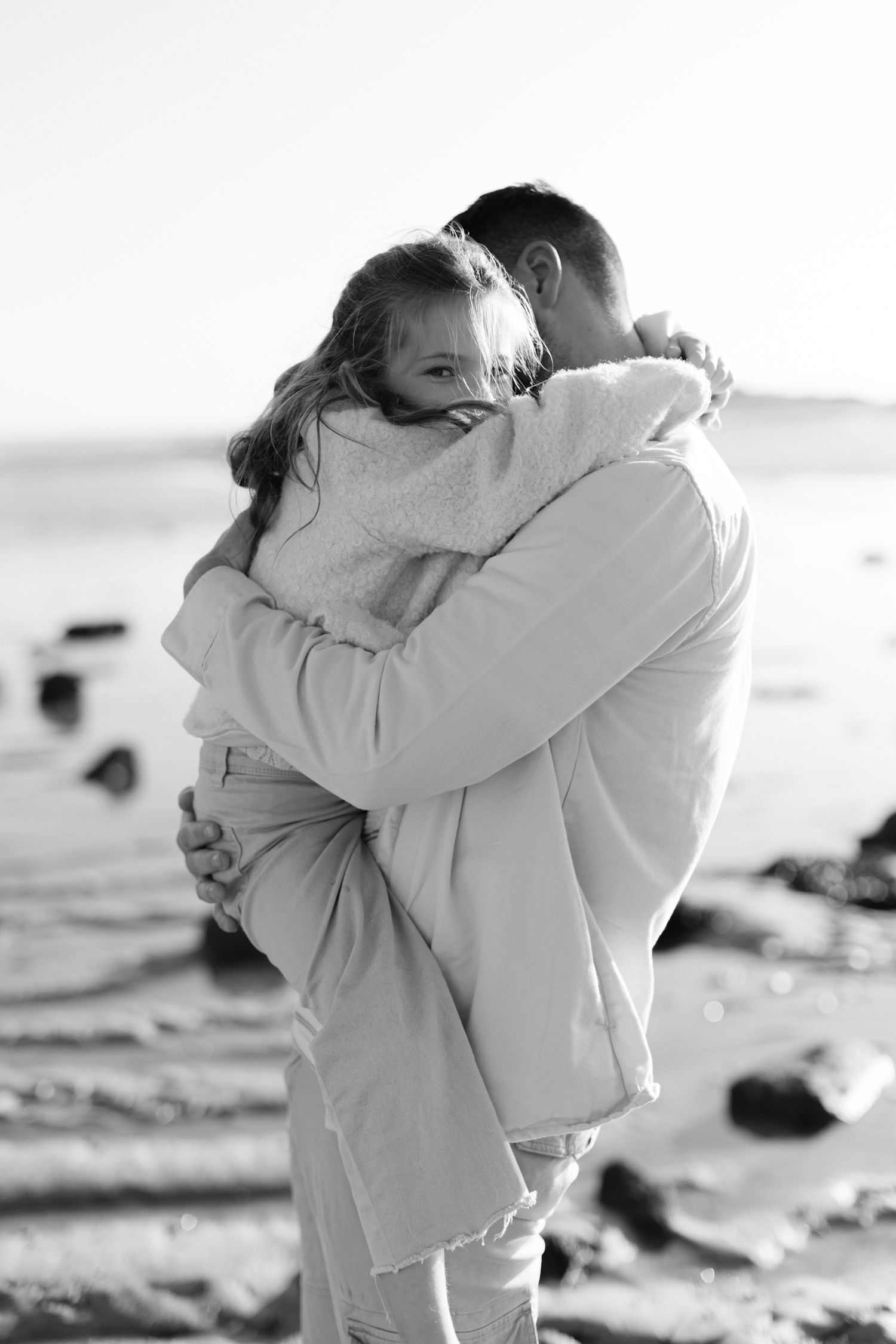 Photo en noir et blanc d'un père tenant et serrant sa jeune fille dans ses bras sur une plage rocheuse.