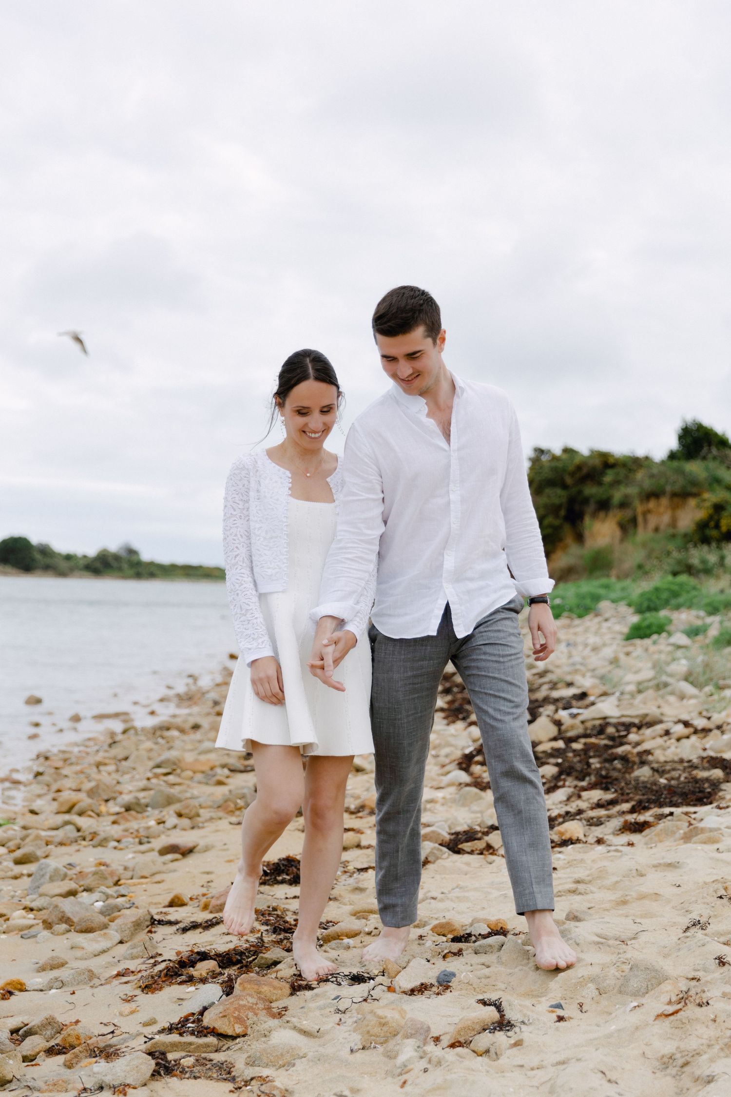 Un couple en tenue blanche marche pieds nus sur une plage rocheuse, souriant et se tenant la main par une journée nuageuse.