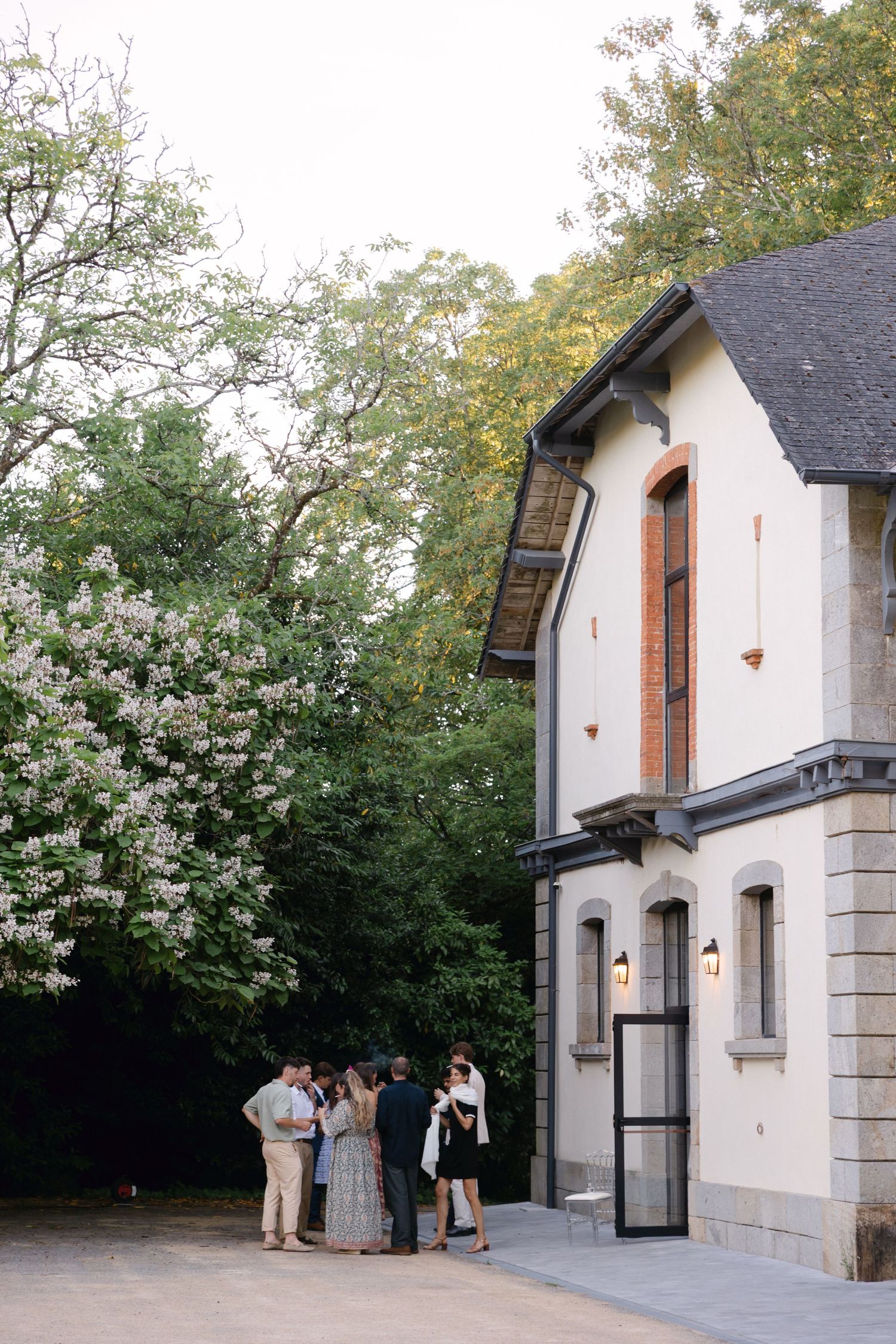 Les invités se rassemblent à l'extérieur d'un élégant bâtiment en pierre blanche aux bordures sombres, entouré d'arbres luxuriants.