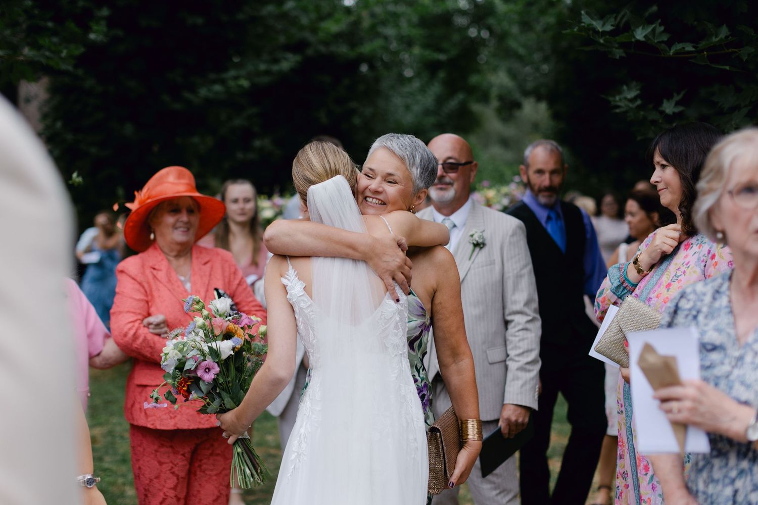 Mariée en robe en dentelle blanche embrassant une invitée plus âgée tout en tenant un bouquet coloré après la cérémonie.