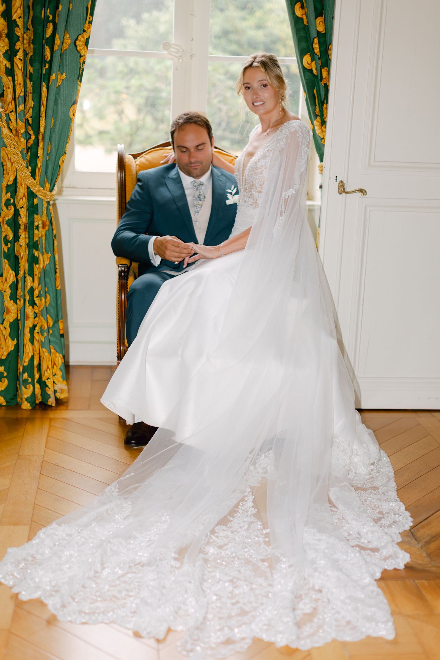 La mariée en robe de mariée en dentelle avec voile de cape se tient à côté du marié assis en costume sarcelle.