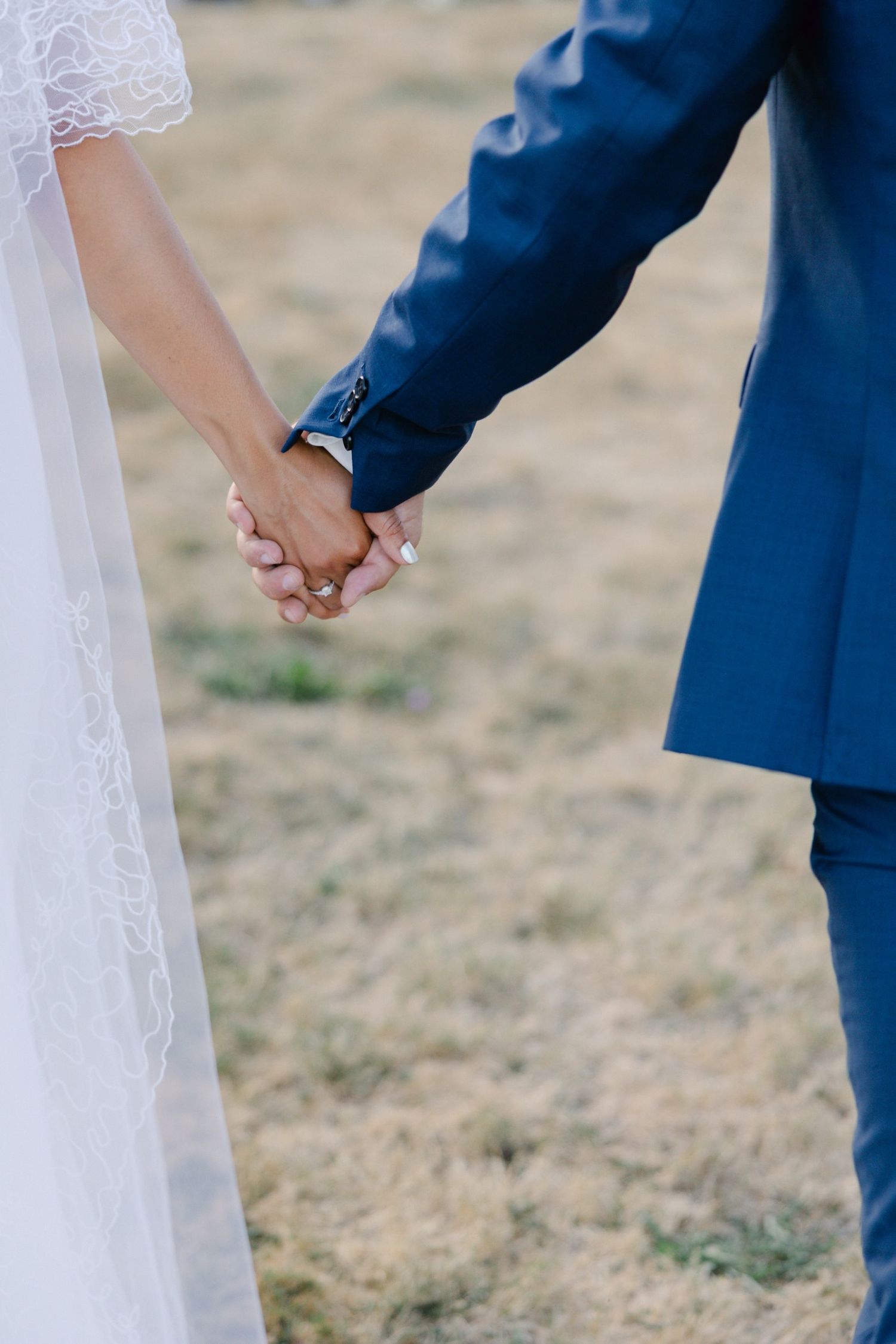 A bride and groom hold hands outdoors, she in a white lace gown, he in a navy blue suit, on their wedding day.