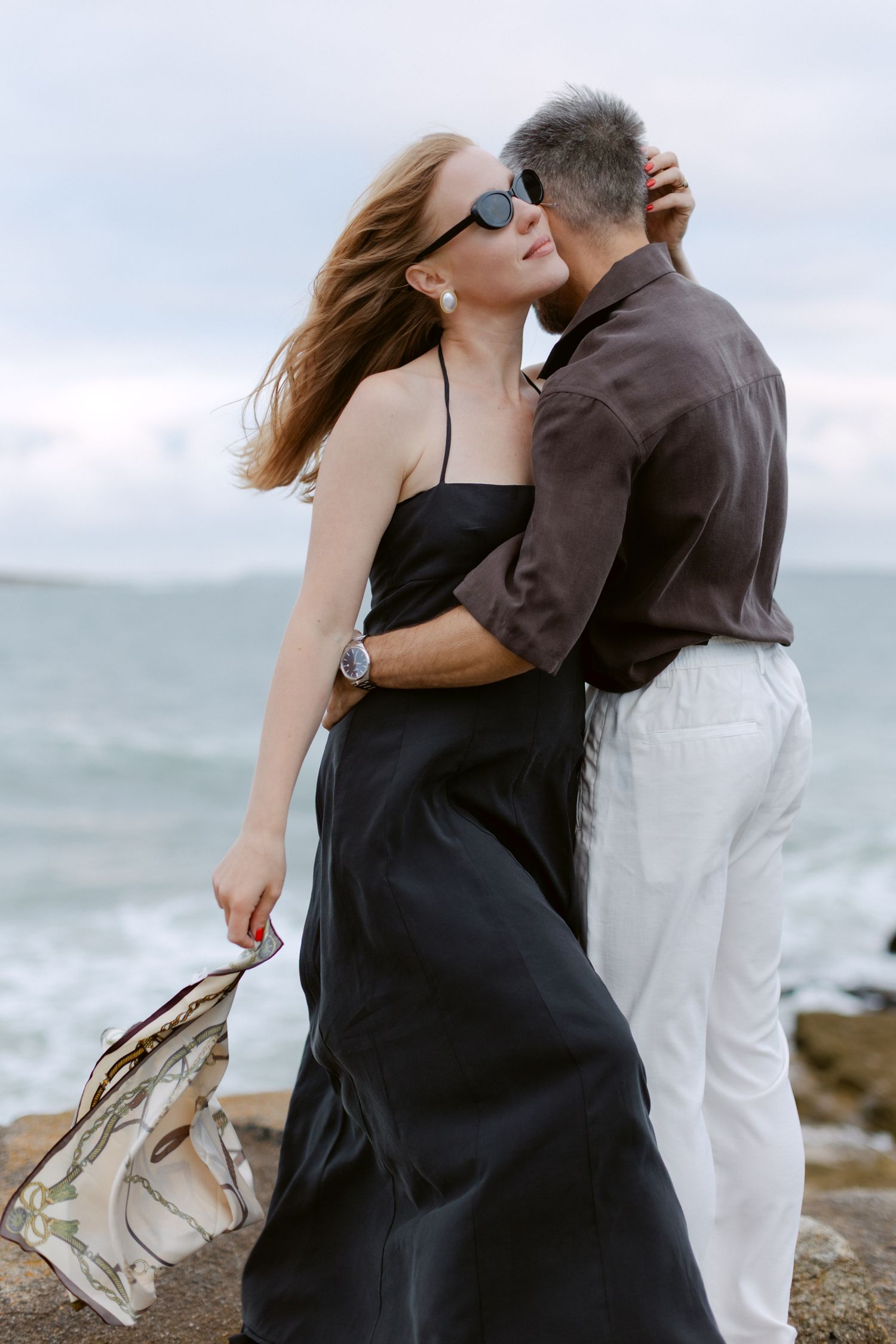 Un couple s'embrasse romantiquement au bord de l'océan, une femme en robe noire aux cheveux coulants, un homme en chemise marron et pantalon