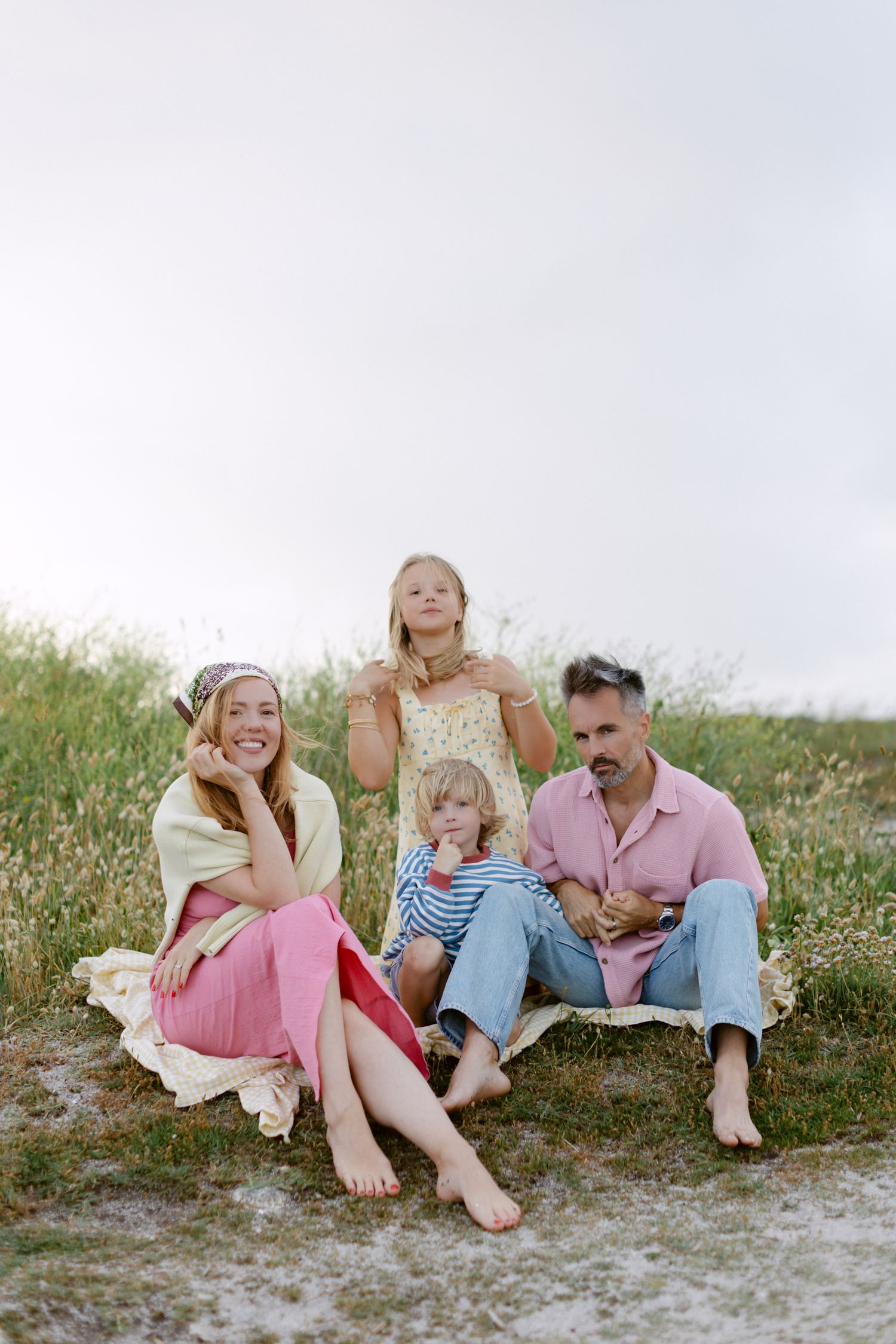 Famille de quatre personnes assise pieds nus sur l'herbe, parents et deux enfants en tenues pastel profitant d'un moment en plein air.
