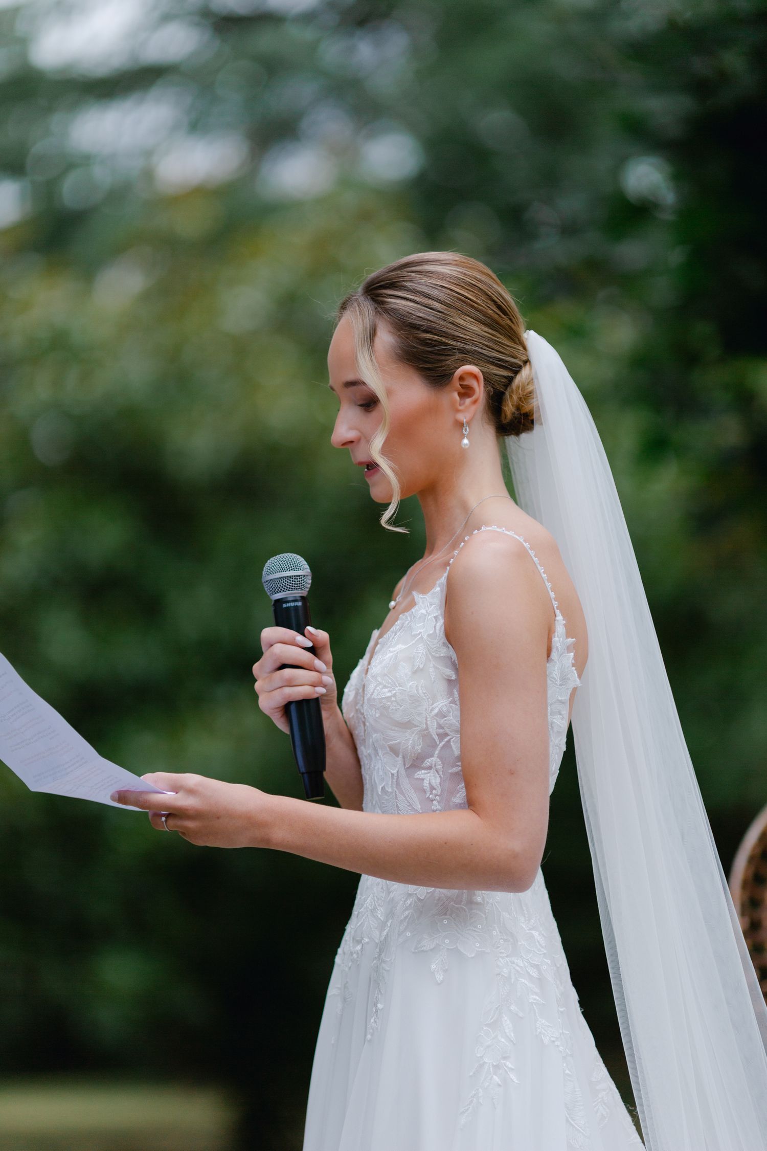La mariée en robe de dentelle et voile lit les vœux de mariage dans un microphone lors d'une cérémonie en plein air.