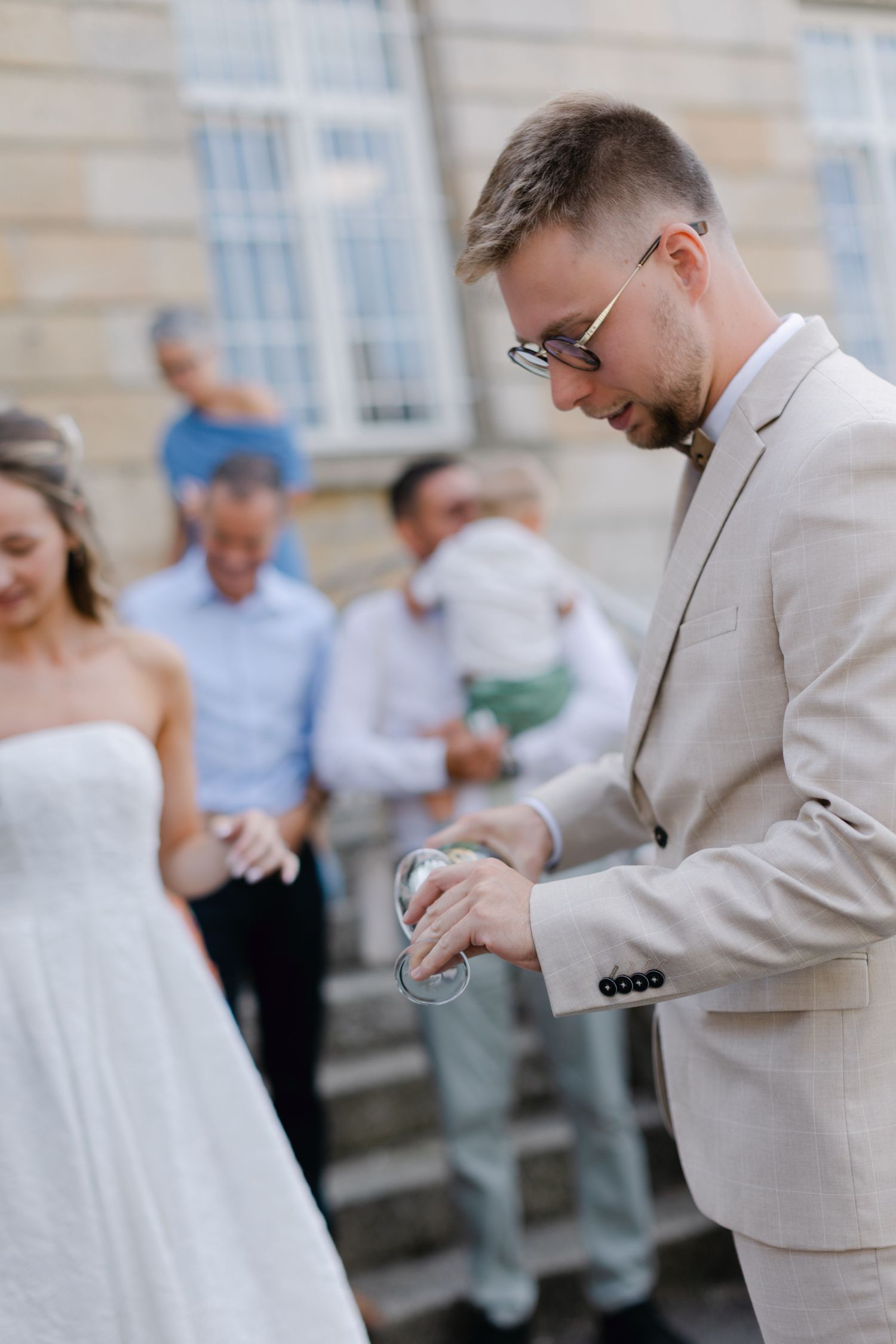 Le marié en costume beige et nœud papillon verse du champagne à côté de la mariée en robe blanche sans bretelles après le mariage.