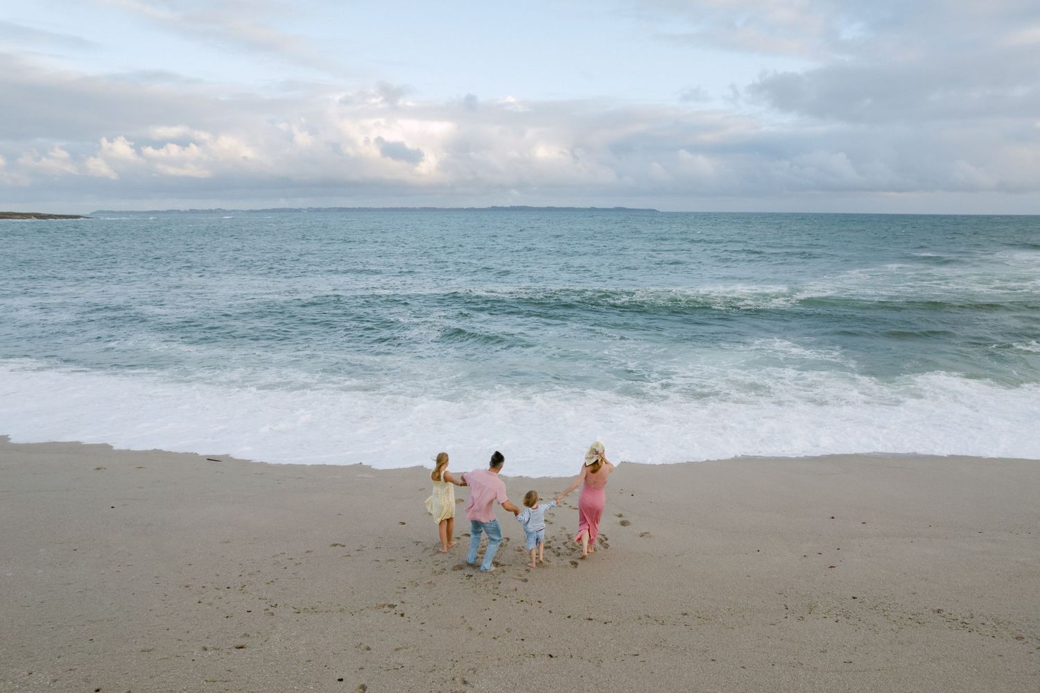 Famille de quatre personnes se tenant la main sur une plage de sable près des vagues de l'océan sous un ciel nuageux.