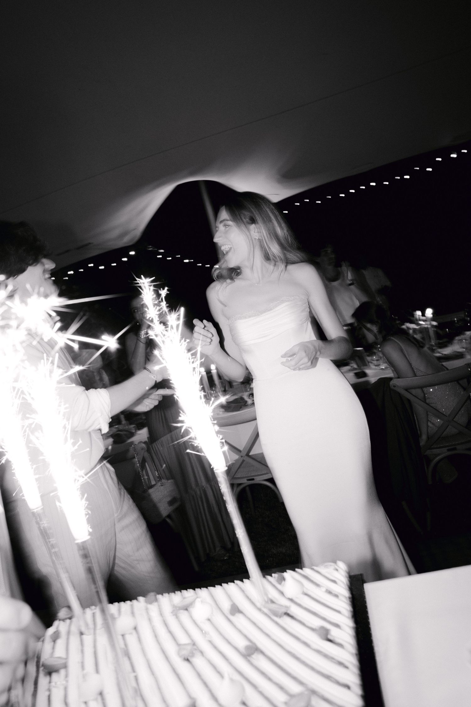 Photo en noir et blanc d'une mariée en robe blanche souriante près d'un gâteau de mariage avec des cierges magiques lors d'une réception.