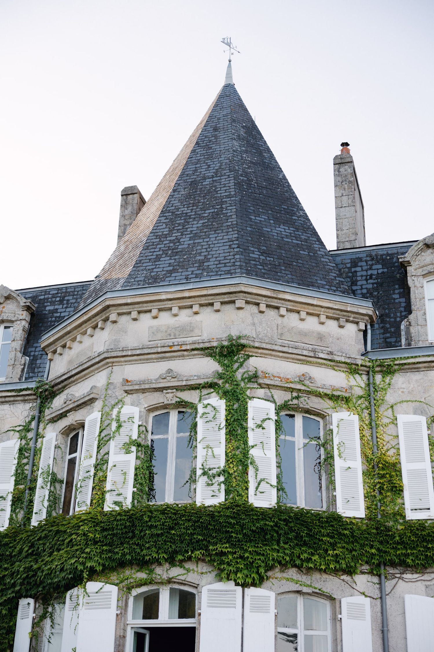 French chateau with ivy-covered stone facade, white shutters, pointed slate turret roof, and weathervane against overcast sky.
