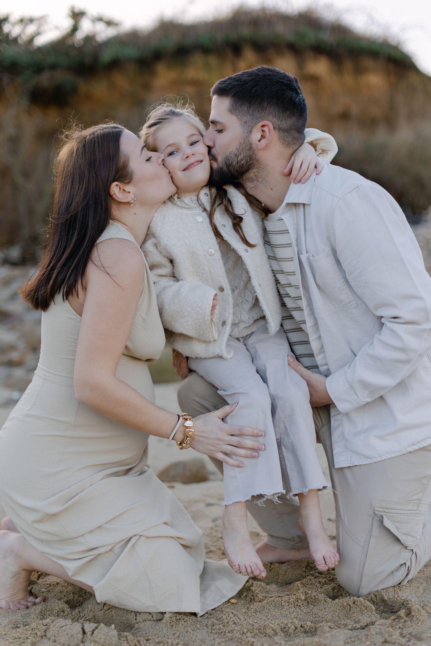 Deux parents embrassent leur jeune fille souriante sur les joues alors qu'ils sont assis sur une plage.