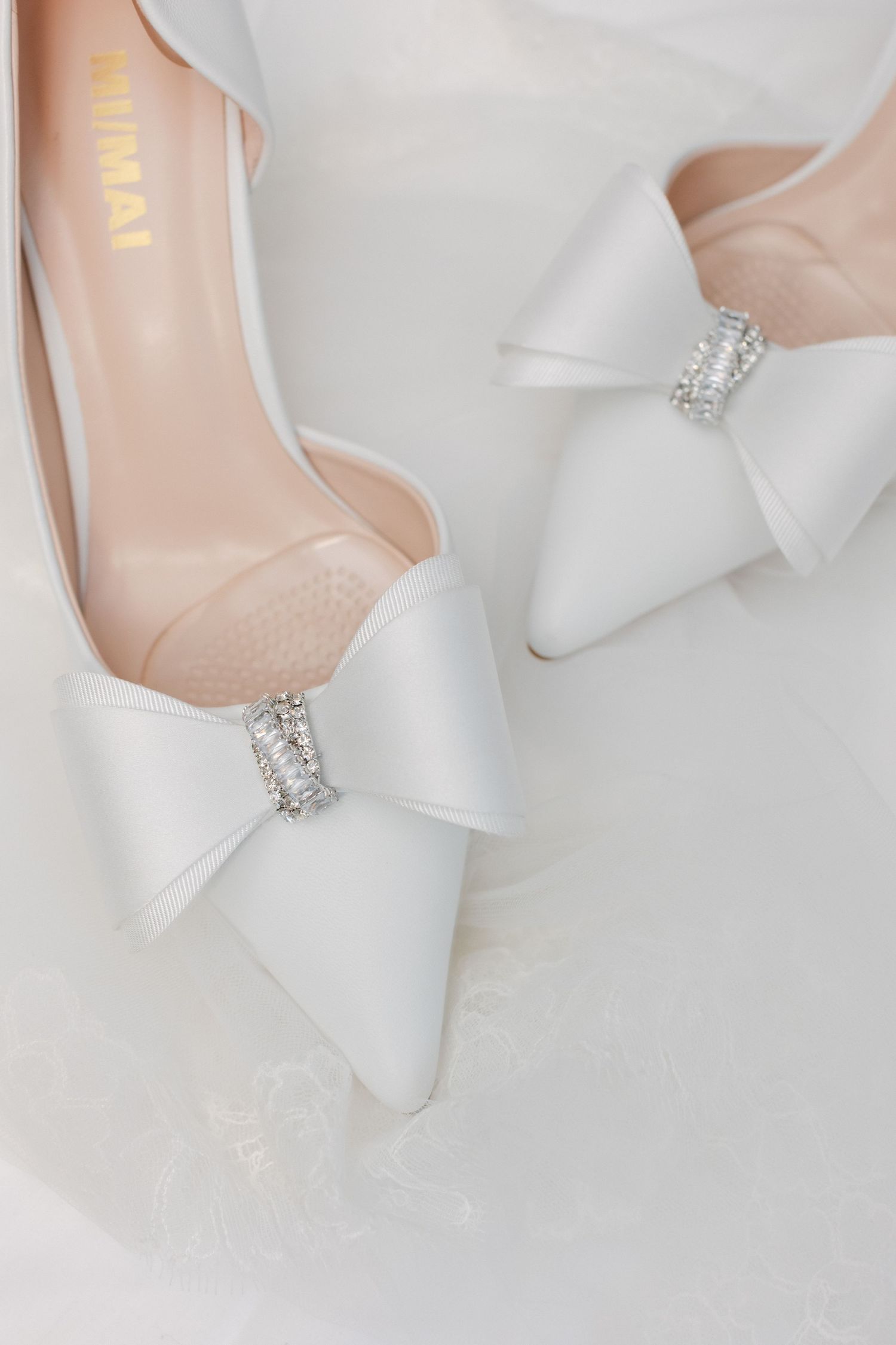 Talons de mariée en satin blanc avec nœud et détail d'embellissement en cristal sur fond blanc doux.