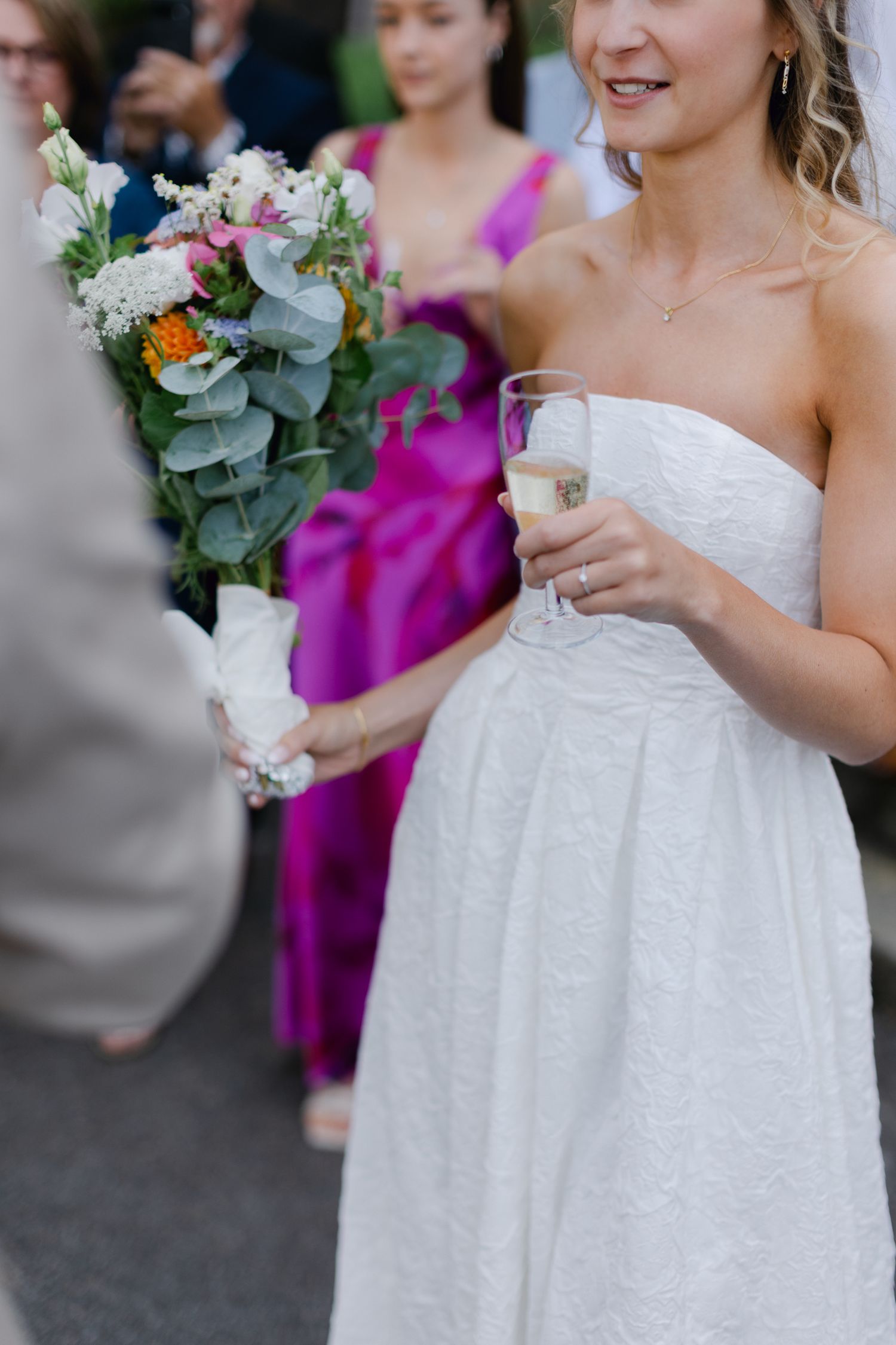 La mariée en robe en dentelle blanche sans bretelles tient une flûte à champagne et un bouquet coloré pendant que les invités célèbrent.