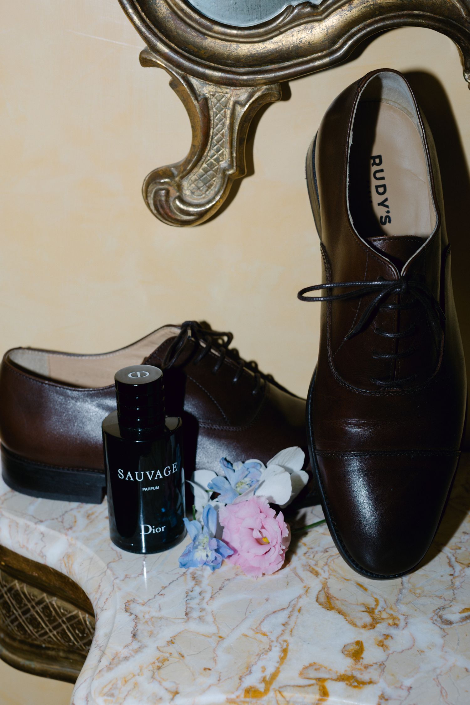 Chaussures Oxford en cuir marron du marié avec eau de Cologne Dior Sauvage et boutonnière florale sur surface en marbre.