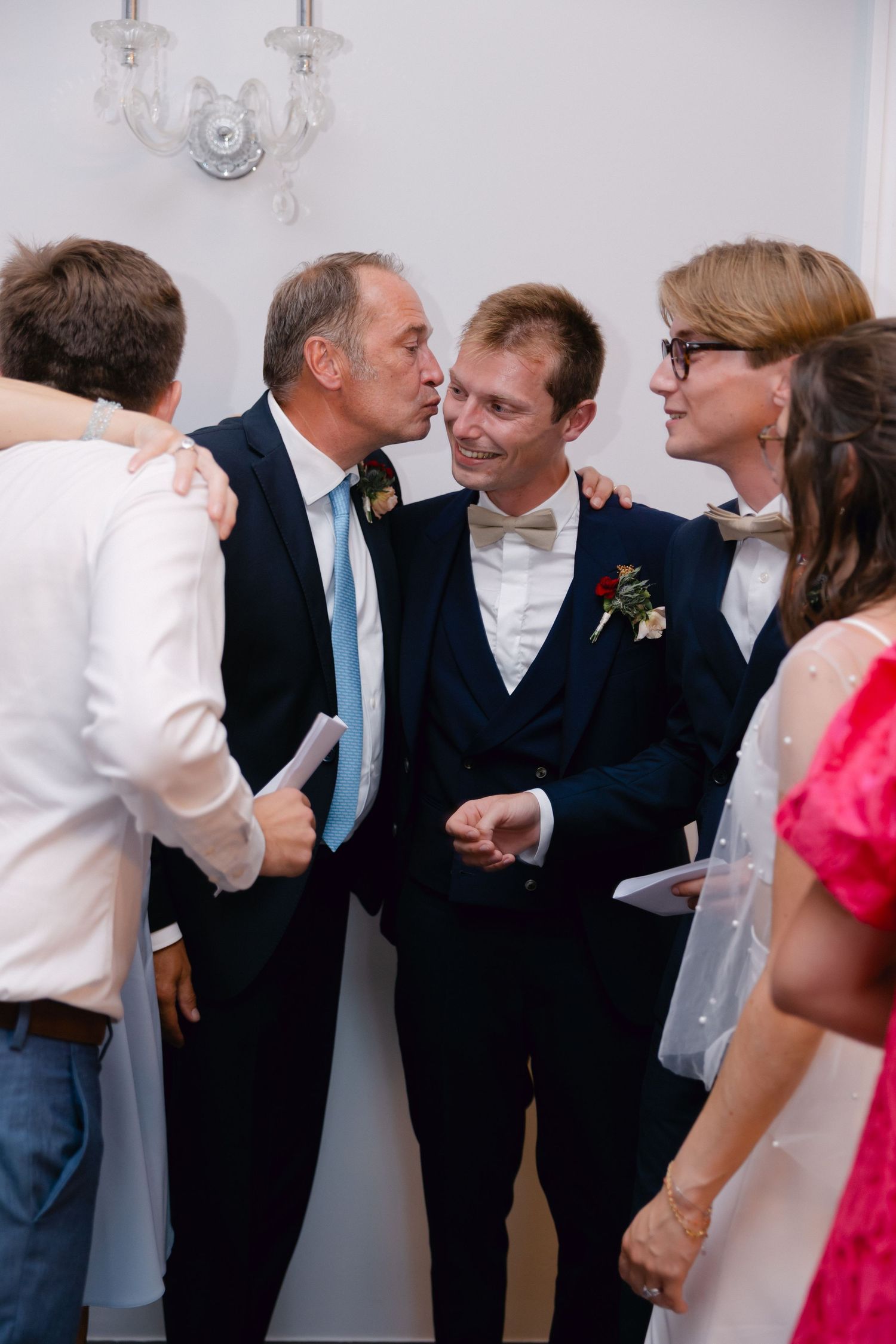Le marié en costume bleu marine reçoit un baiser sur la joue d'un homme plus âgé lors d'une réception de mariage avec des invités.