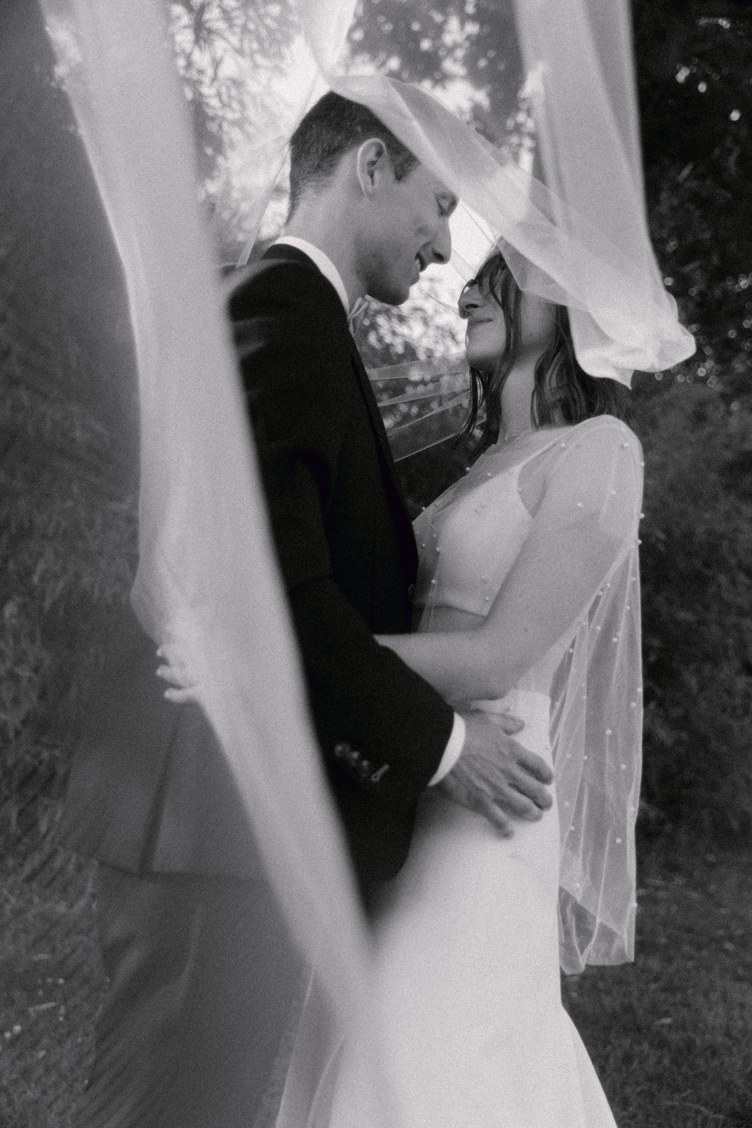 Photo en noir et blanc de la mariée et du marié s'embrassant sous un voile fluide à l'extérieur le jour de leur mariage.